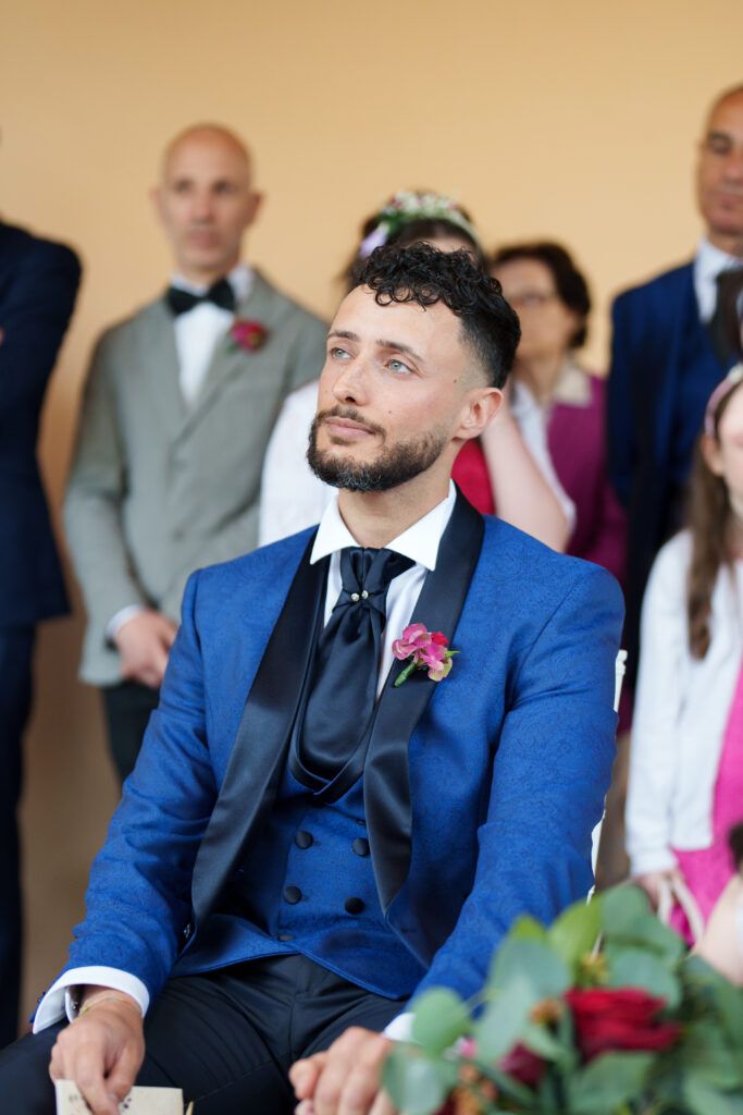 Wedding guest in blue three-piece suit with pink boutonniere seated during ceremony.
