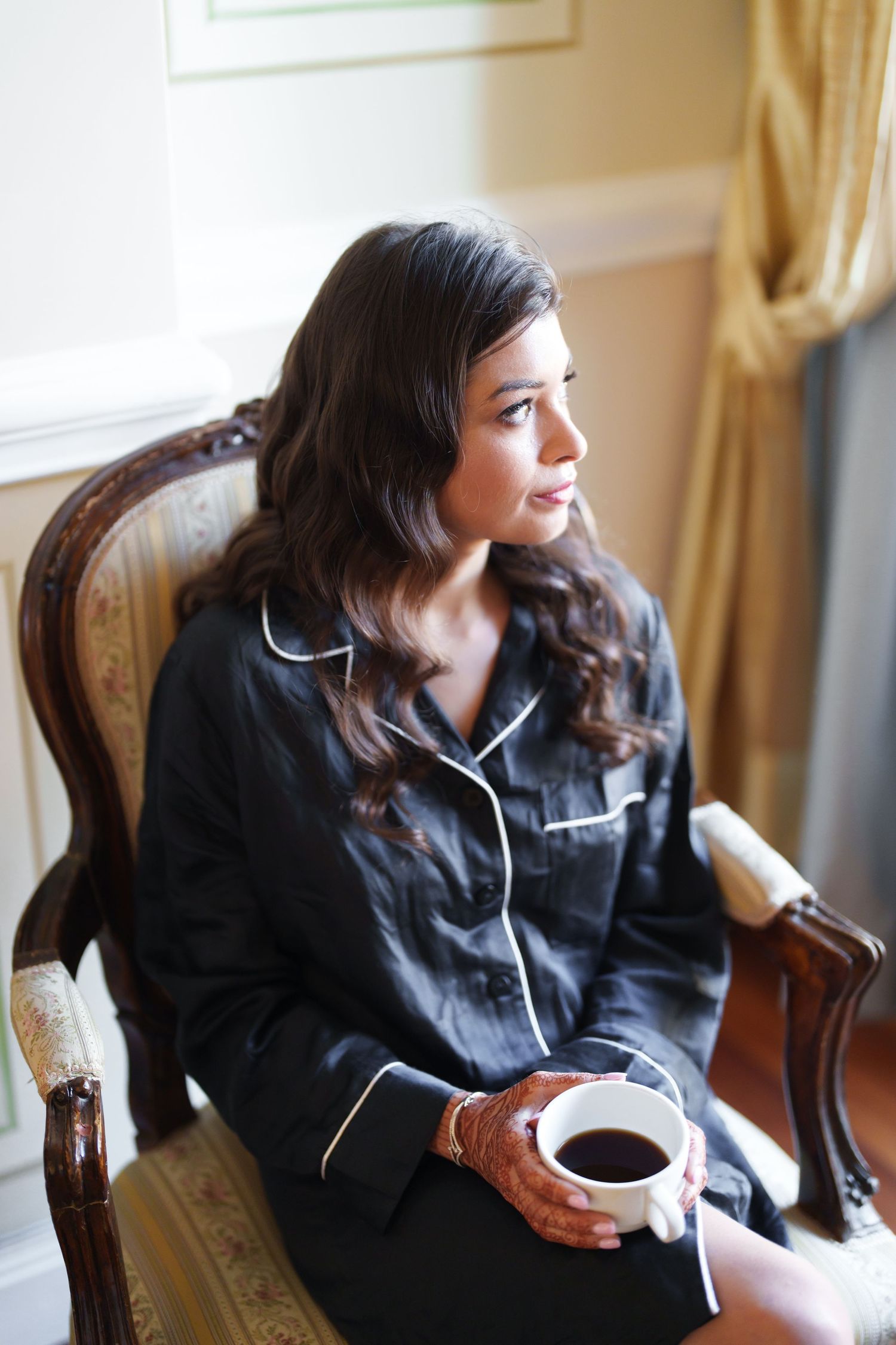 Person relaxing in black sleepwear on an antique chair by a window in an elegant room.