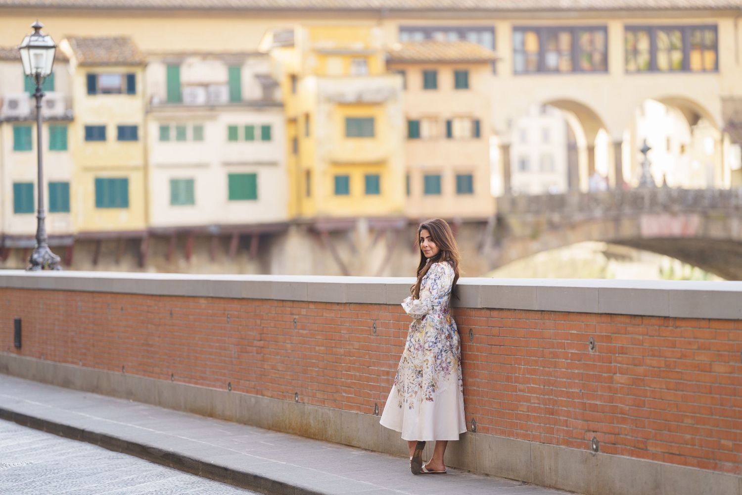 Woman in floral dress posing by brick wall with Ponte Vecchio bridge in Florence, Italy in background.