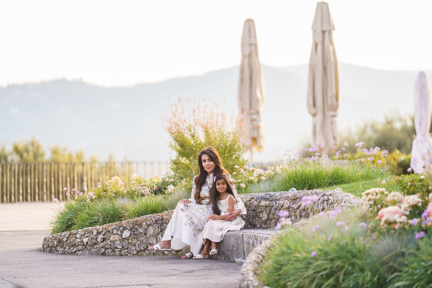 A mother and daughter sit together on stone steps surrounded by lush gardens and mountains in the background.