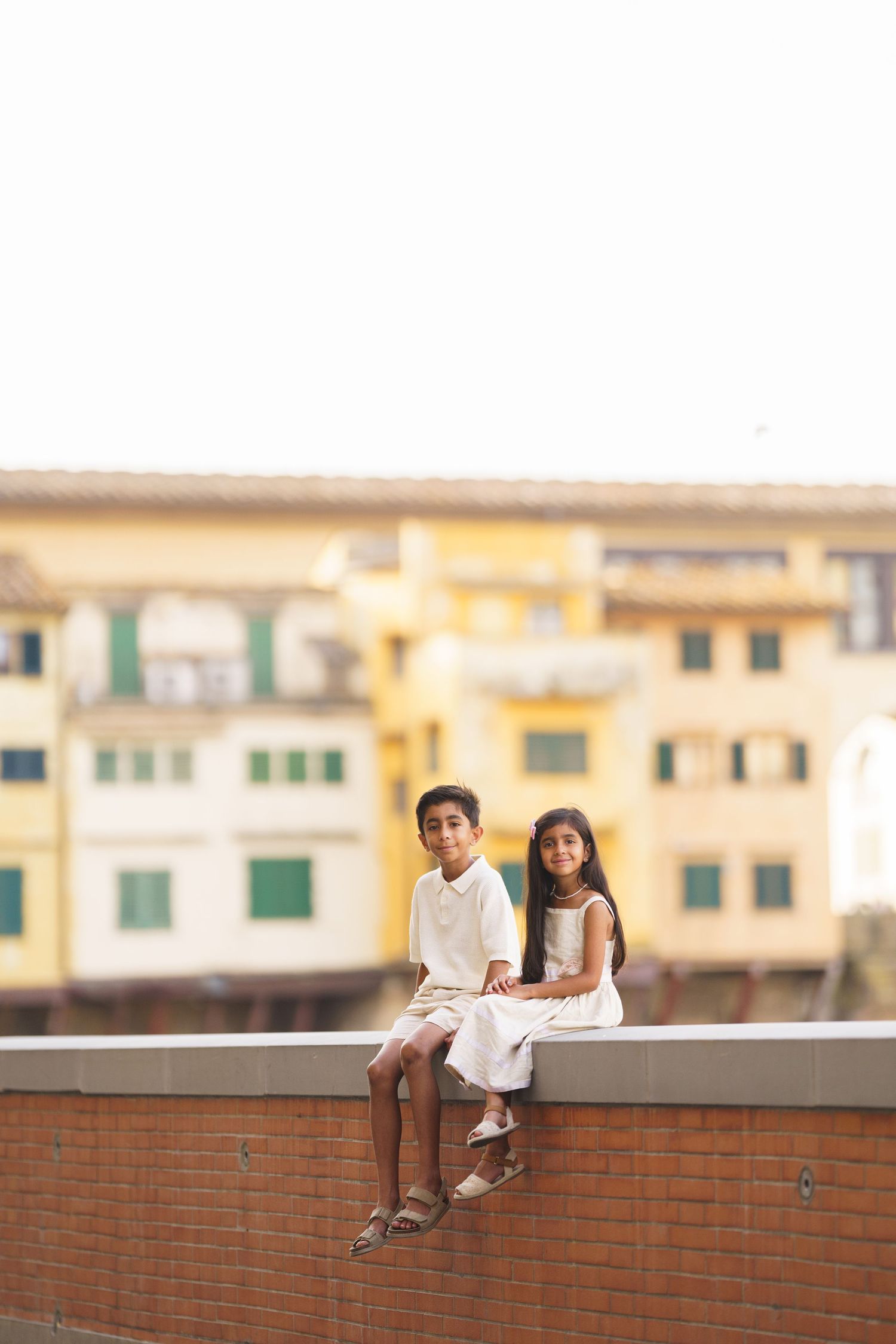 Two children in white outfits sitting on a wall with colorful Italian buildings in the background.