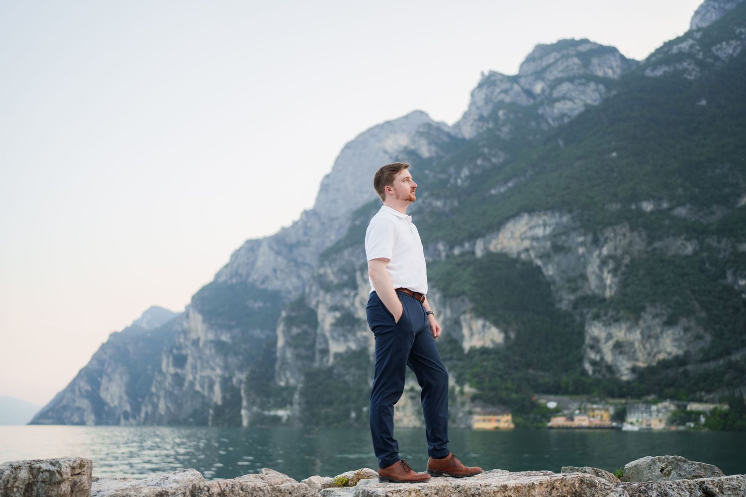 Stylish man in white shirt and navy pants standing confidently on rocks before mountain lake.