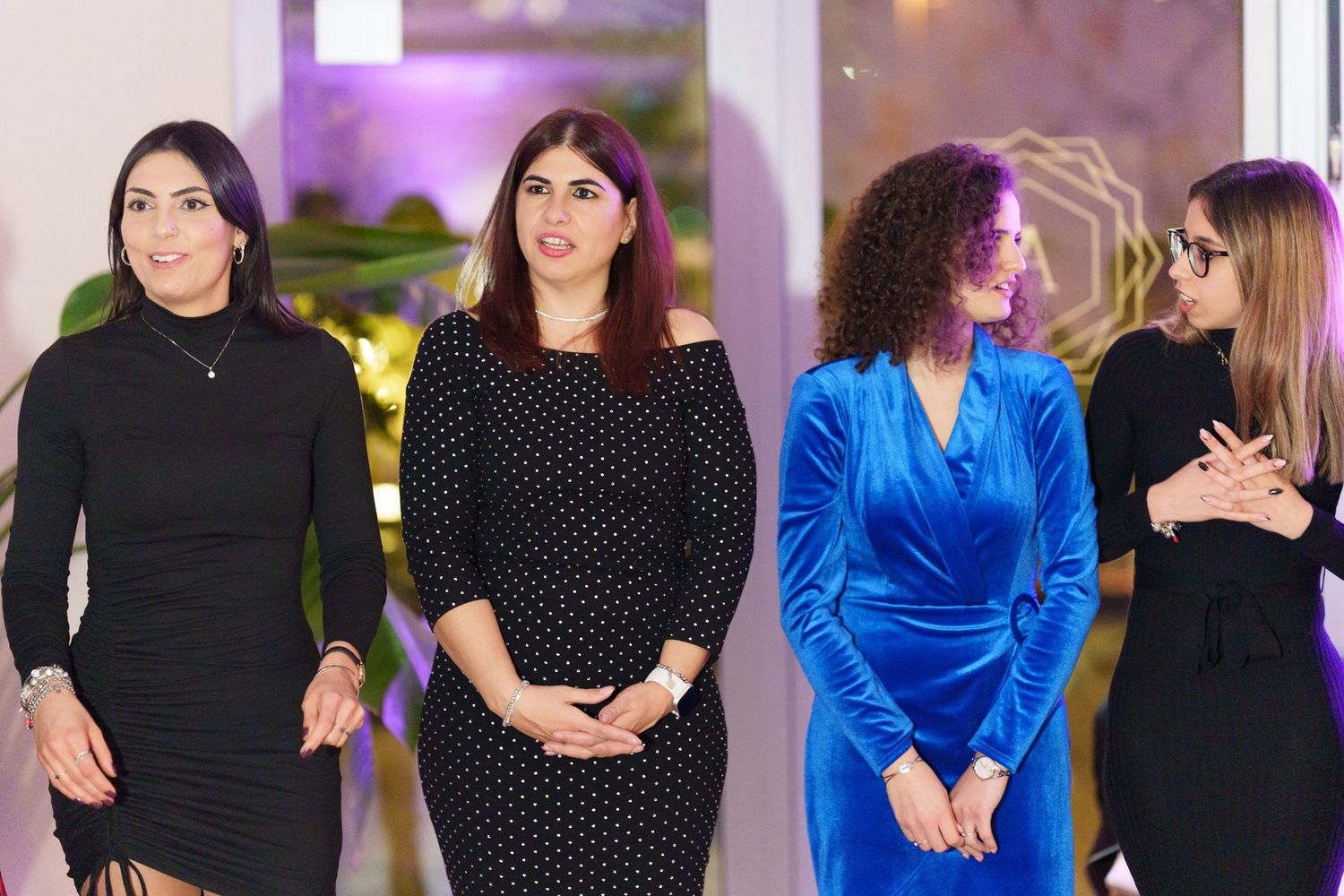 Four women standing together at an elegant event, wearing stylish black and blue velvet dresses in a decorated venue.