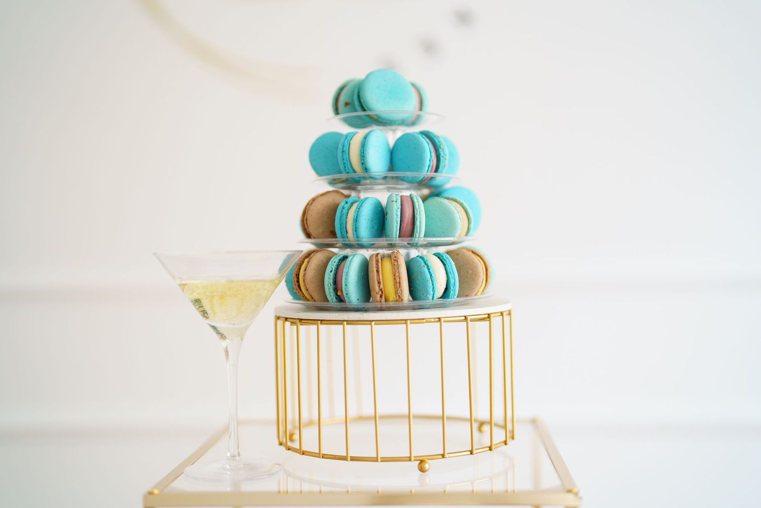 Elegant macaron tower in teal and brown on a gold stand with a champagne glass beside it.