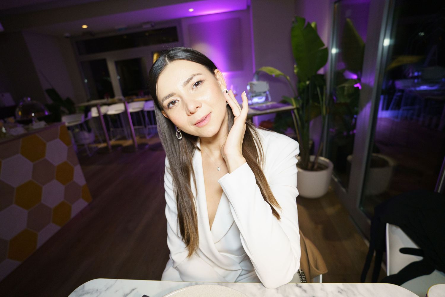 Young woman in white blazer posing elegantly at a restaurant table with purple ambient lighting.
