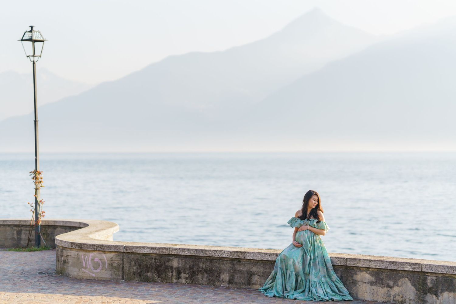 Woman in teal gown sits on stone wall by misty lake with mountain backdrop in serene outdoor portrait.