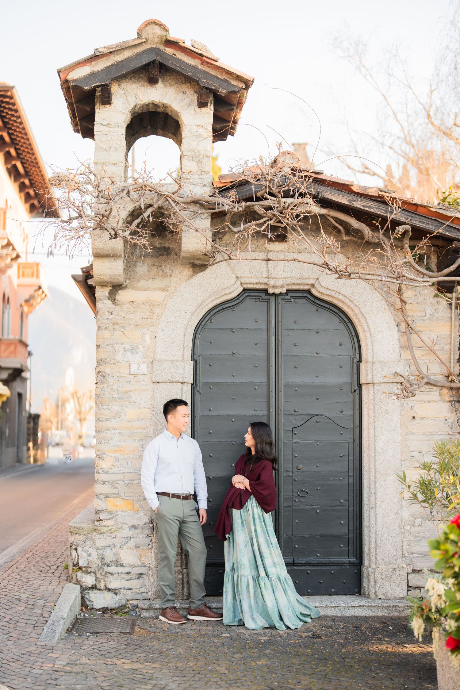 Couple standing before a large arched stone doorway in a charming European street, woman in teal skirt and burgundy shawl.
