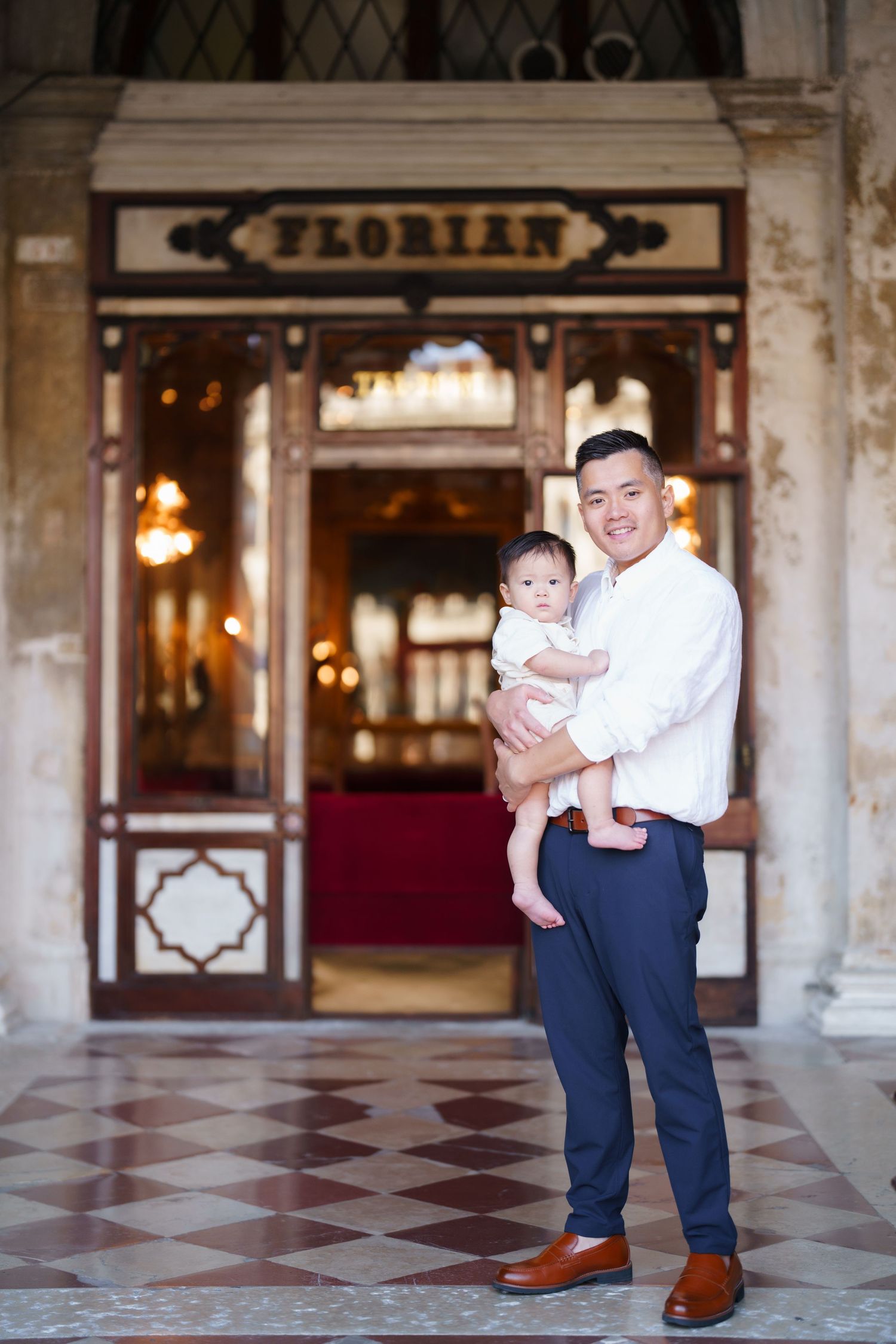 Father holding baby in front of Cafe Florian in Venice, Italy, smiling in elegant attire on checkered marble floor.
