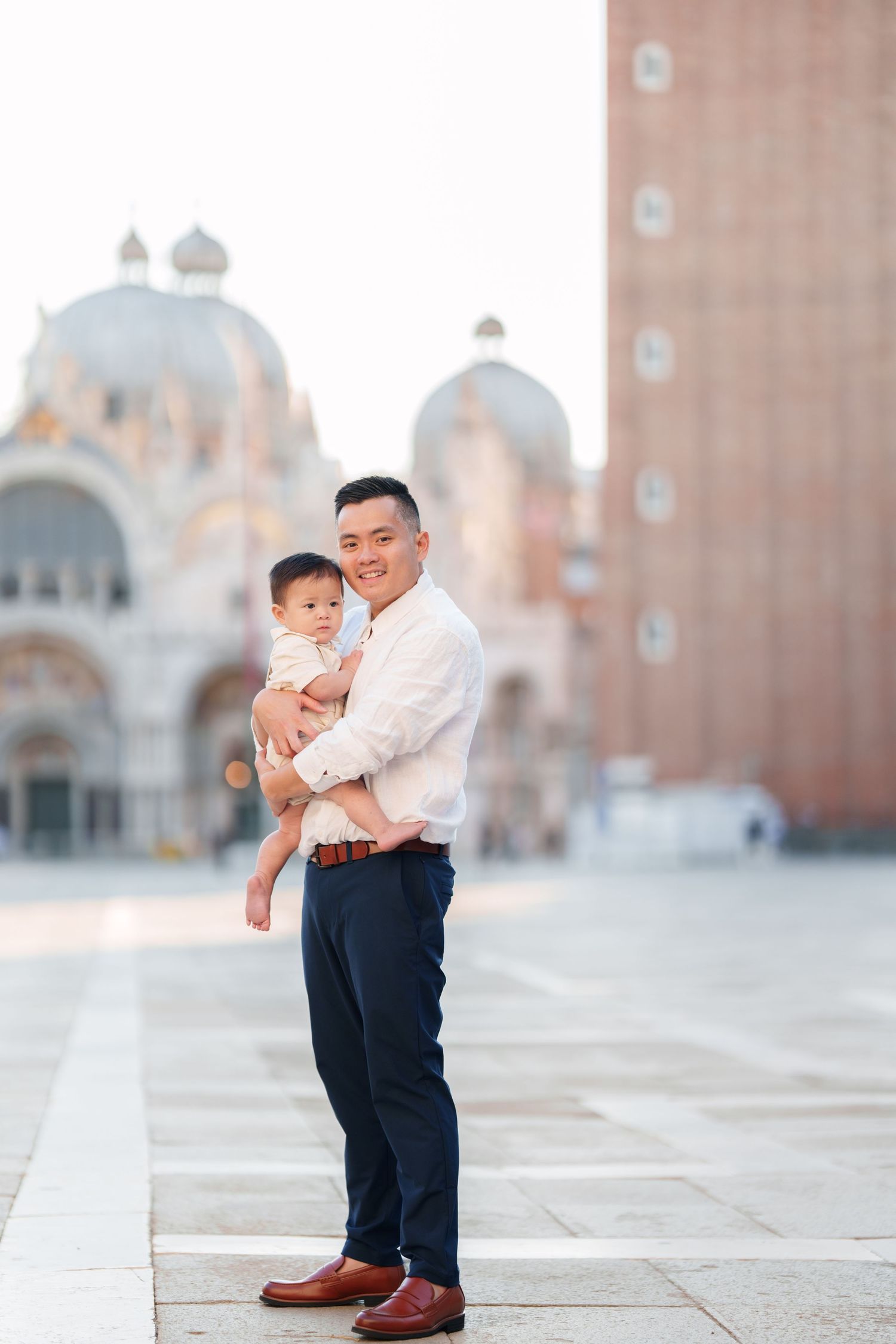 Father holding baby in St. Mark's Square, Venice, with Basilica di San Marco in the background.
