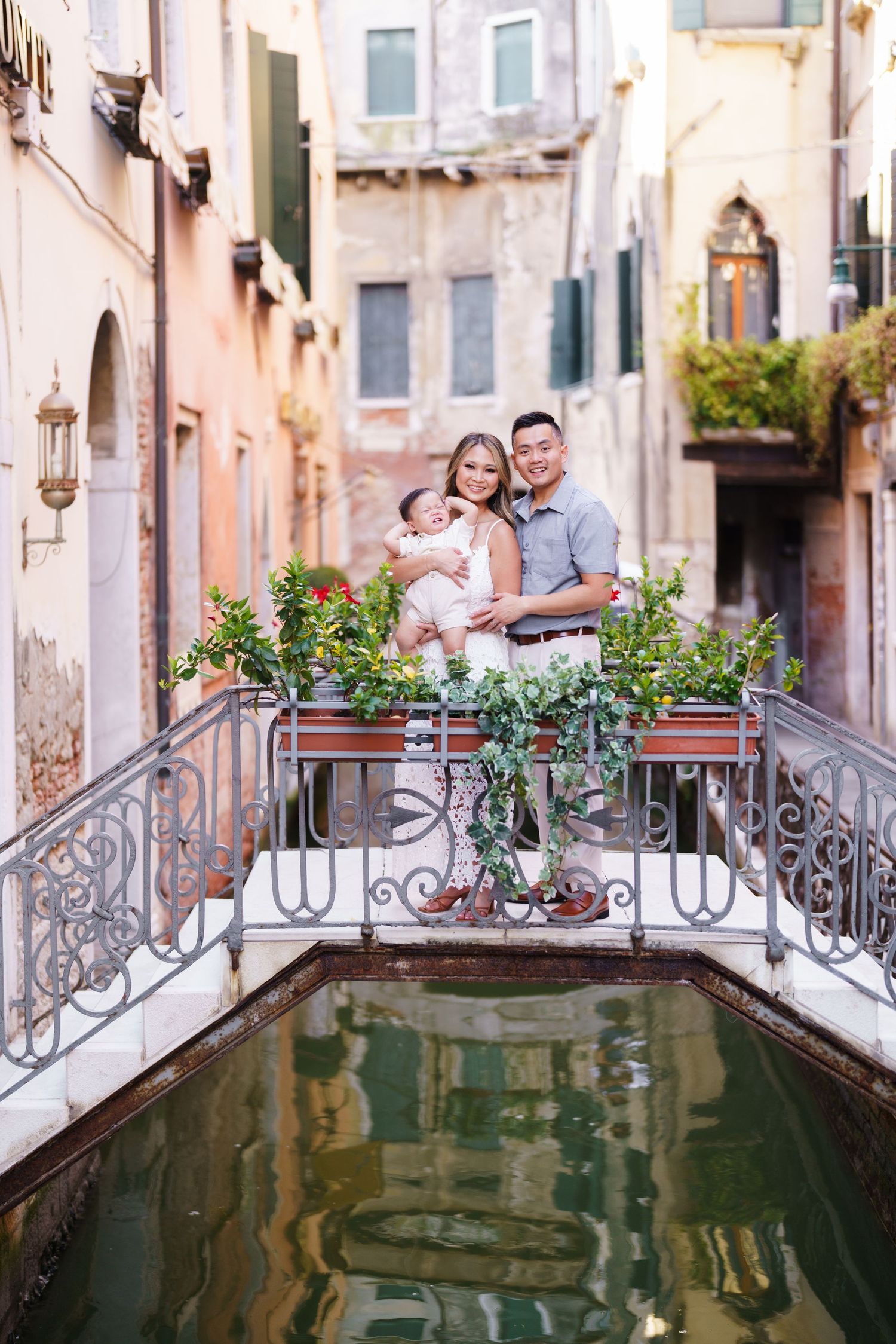 Family of three posing on a charming Venice canal bridge surrounded by lush flowers and historic buildings.