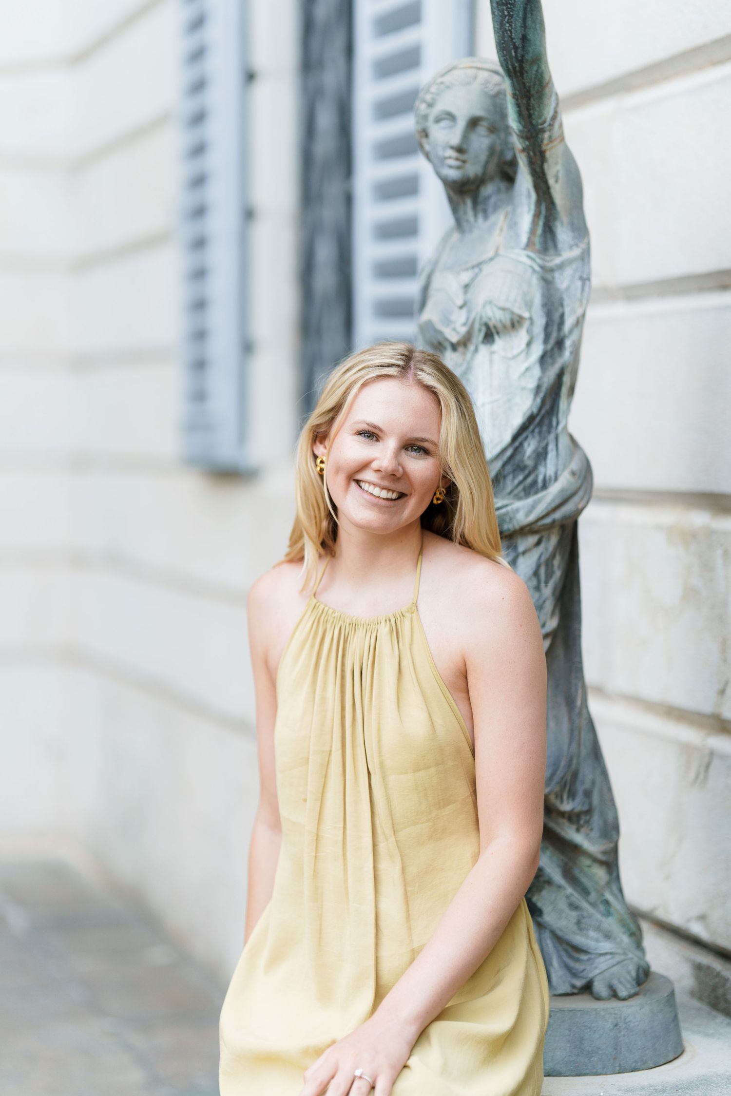 Smiling blonde woman in yellow sundress posing beside a classical stone statue outside a historic building.