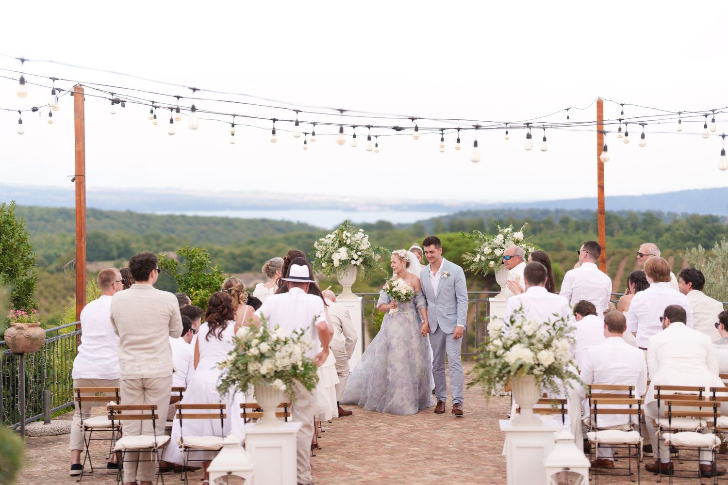 Outdoor wedding ceremony with white floral arrangements, string lights, and scenic vineyard views.