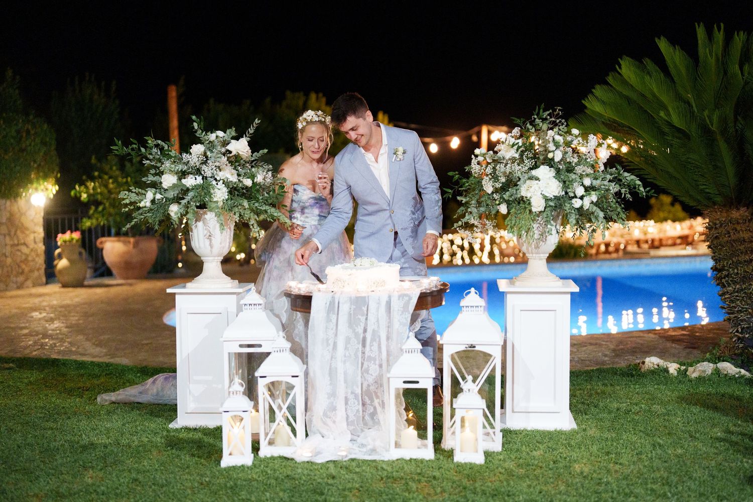 Couple cuts wedding cake at elegant outdoor night reception with white lanterns and floral arrangements by pool.