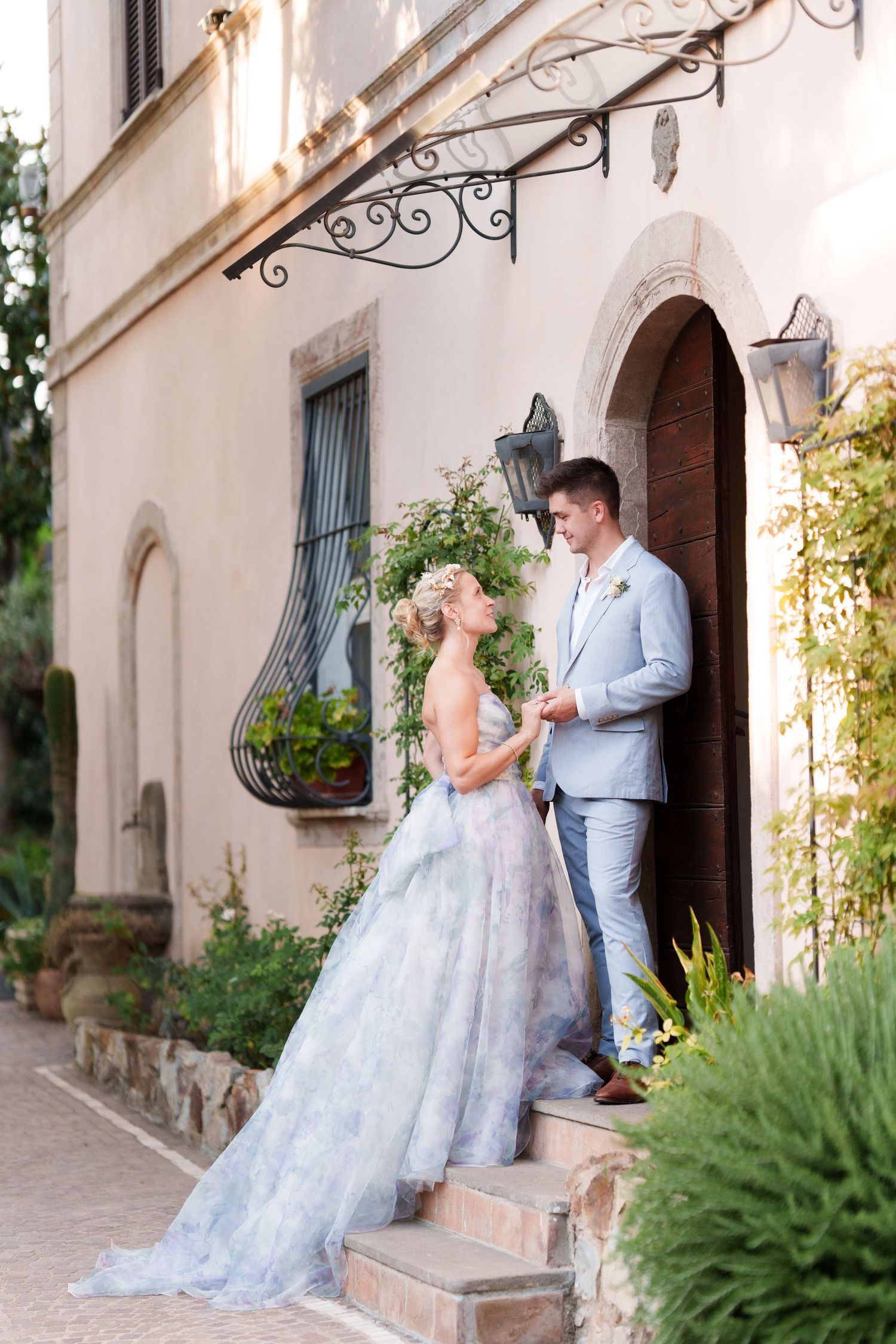 A bride in a floral gown and groom in a blue suit share a romantic moment on stone steps outside a villa.