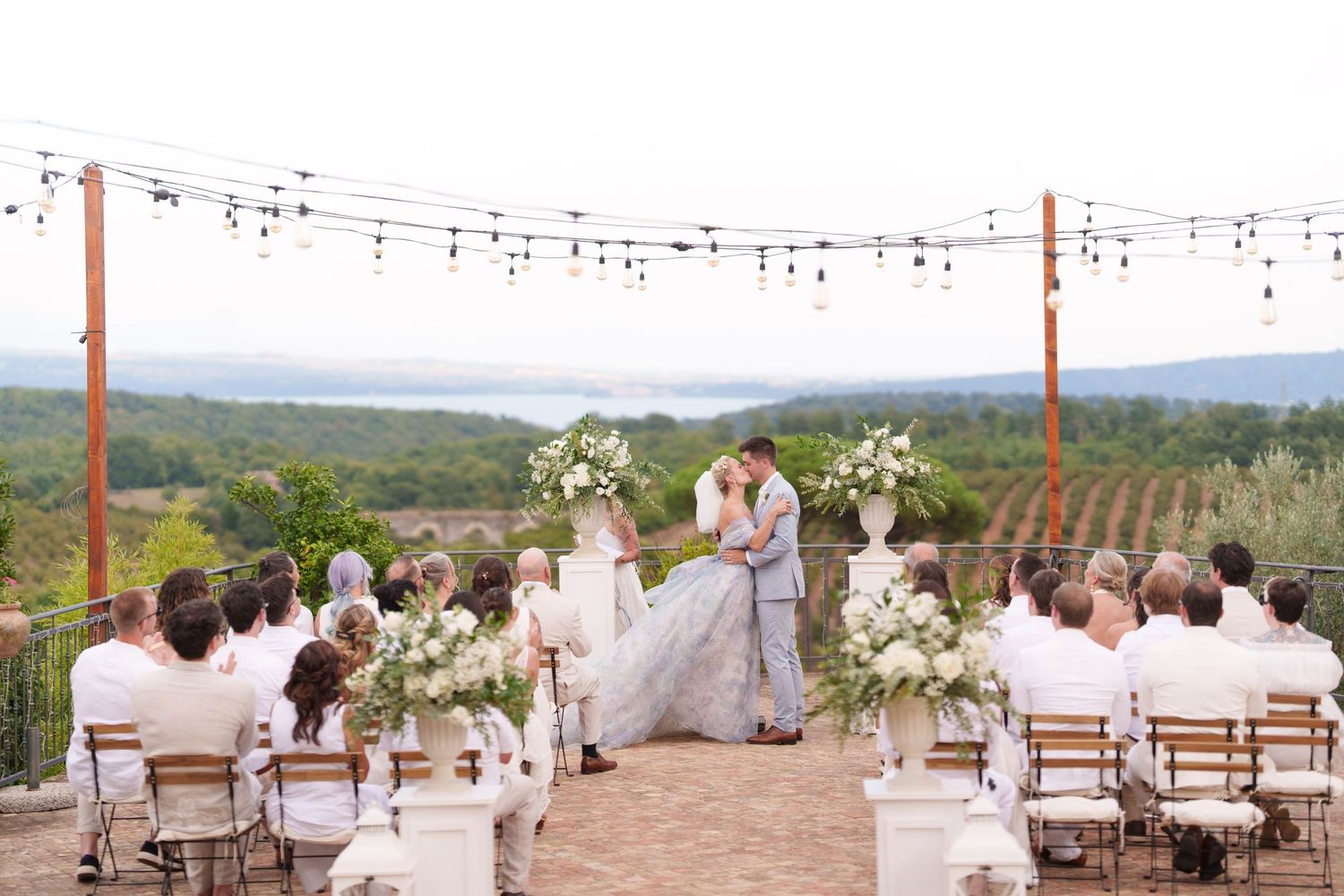 Outdoor vineyard wedding ceremony with couple kissing at altar, white floral arrangements, string lights, and scenic landscape backdrop.