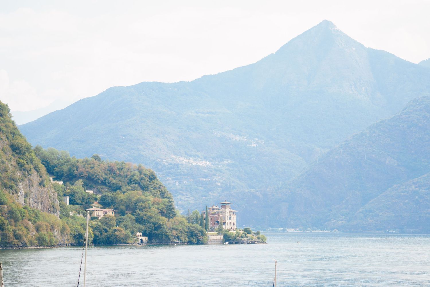 Scenic view of a calm lake surrounded by lush green mountains and a historic villa on the rocky shoreline.