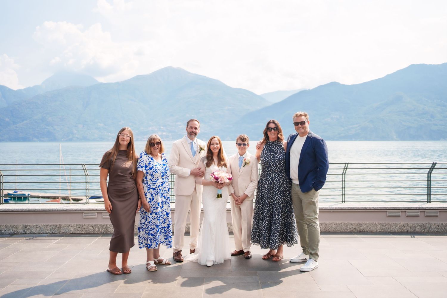 Wedding party poses on lakeside terrace with mountain backdrop on sunny day.