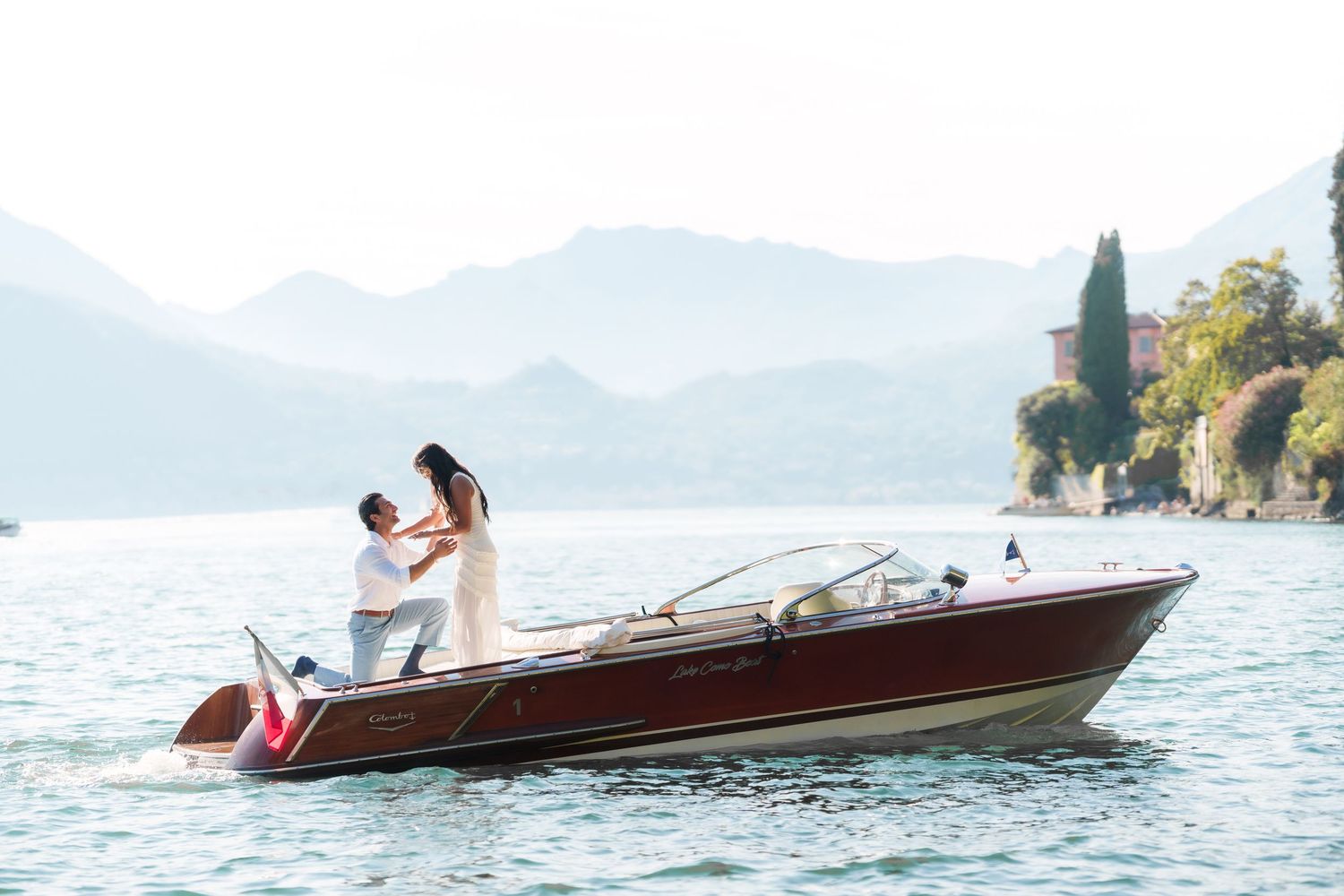 Man proposing to woman on a classic wooden speedboat on a calm lake with misty mountains in the background.