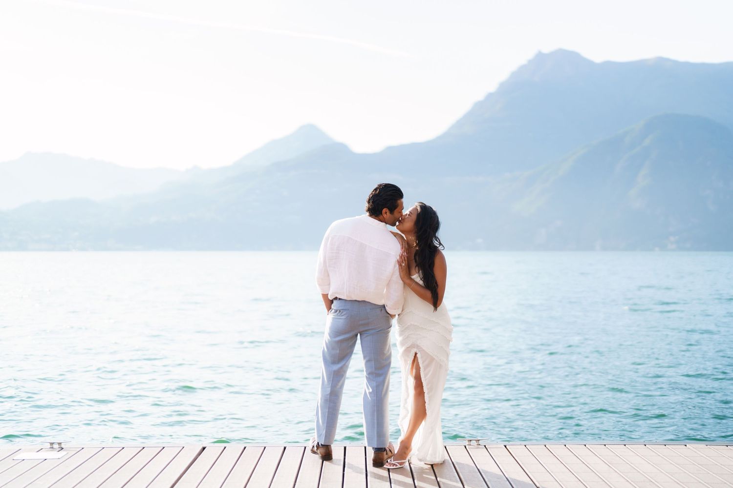 Couple sharing a romantic kiss on a wooden dock overlooking turquoise lake with misty mountain backdrop.