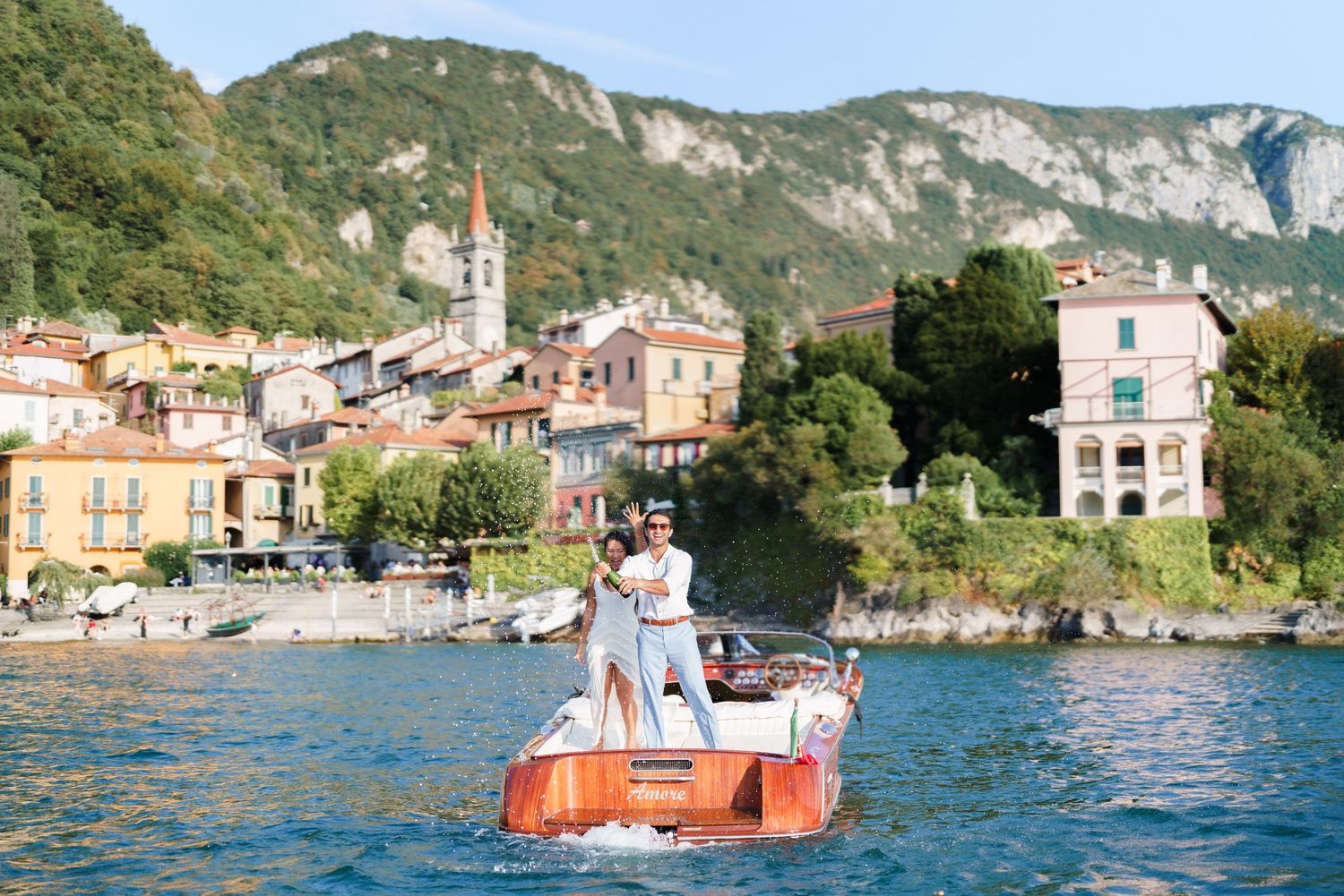 Couple standing on a classic wooden boat on Lake Como with colorful Italian village and mountains in background.