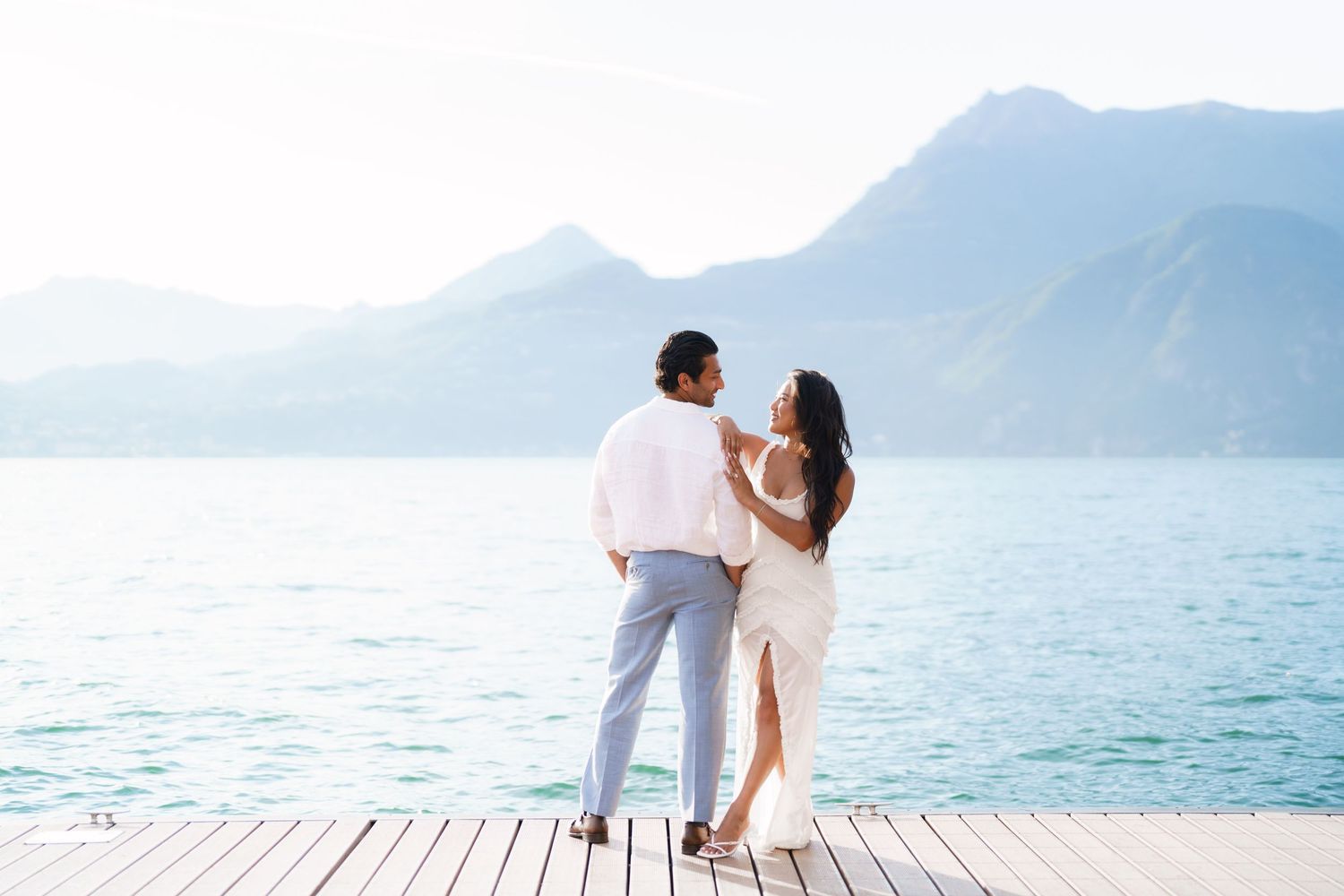 Couple in white outfits standing on a dock overlooking turquoise lake with mountain backdrop.
