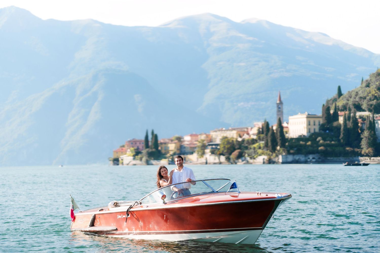 Couple riding a classic red wooden boat on Lake Como with scenic Italian village and mountains in background.