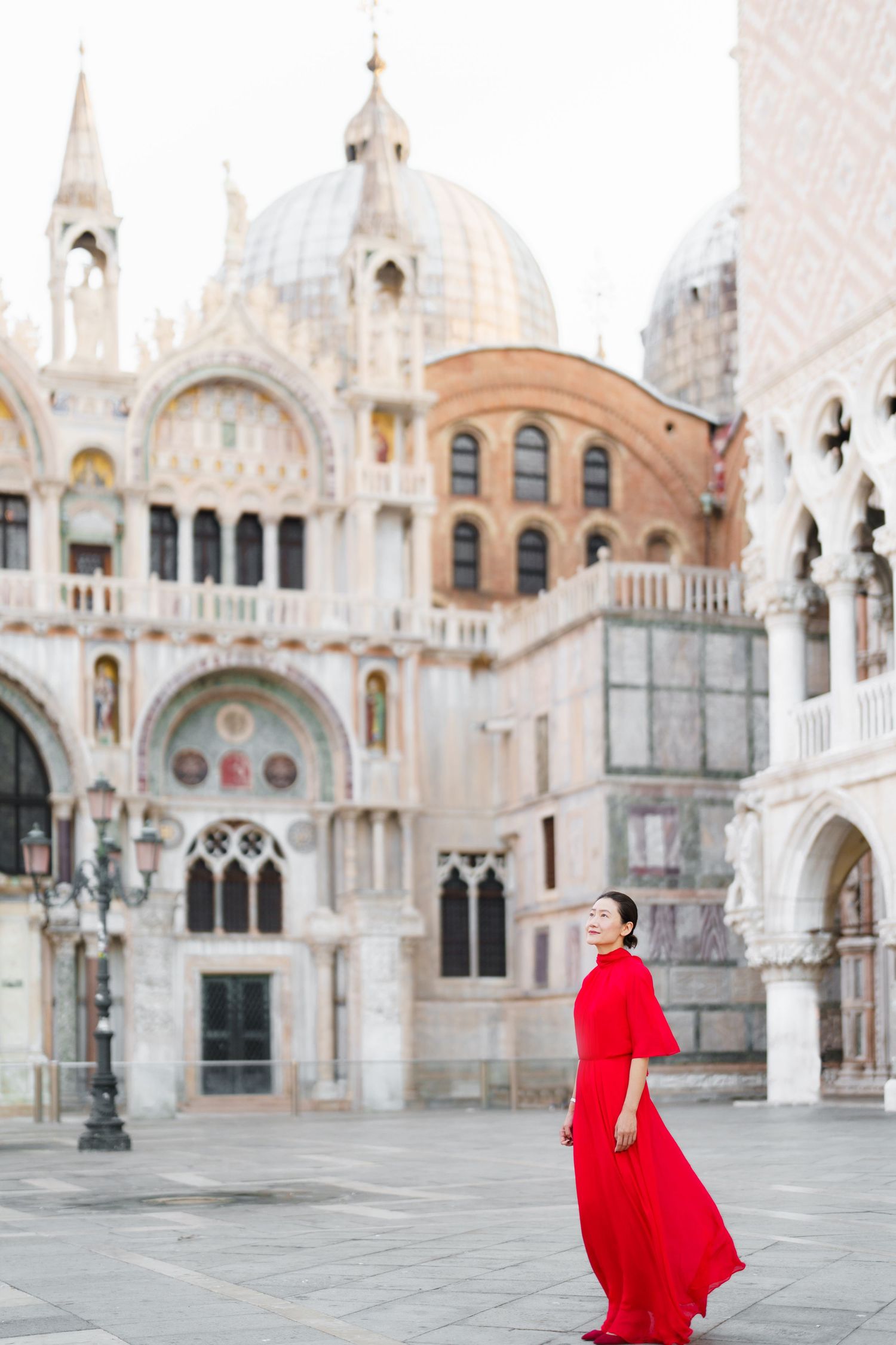 Woman in red dress poses in St. Mark's Square, Venice, with the ornate Basilica facade in the background.