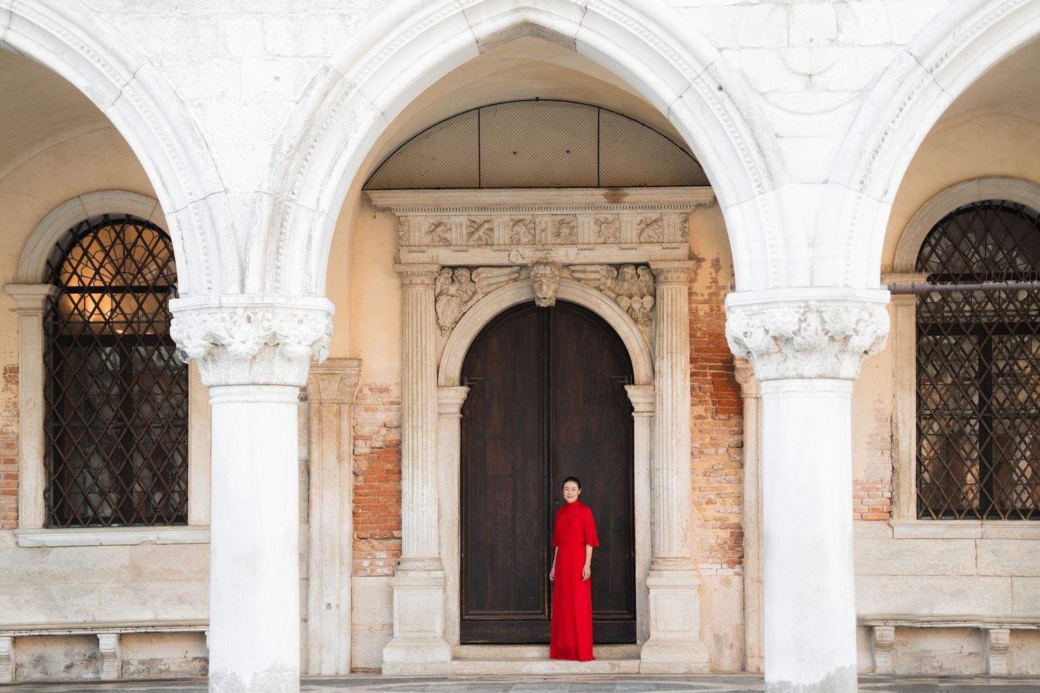 Woman in red gown standing in arched doorway of historic Venetian building with marble columns.