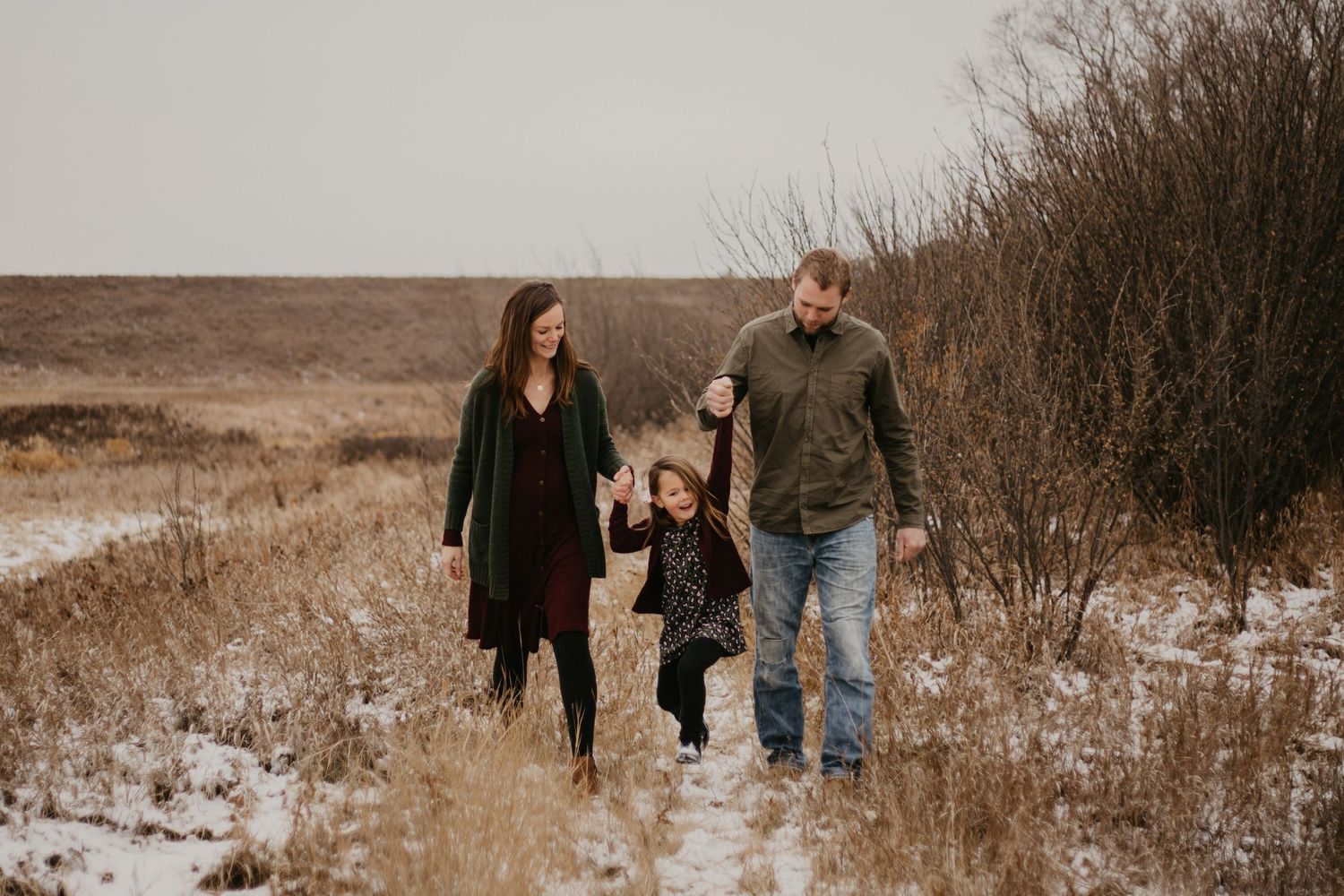 A winter family stroll through a snowy field with brown grasses and shrubs in the background.