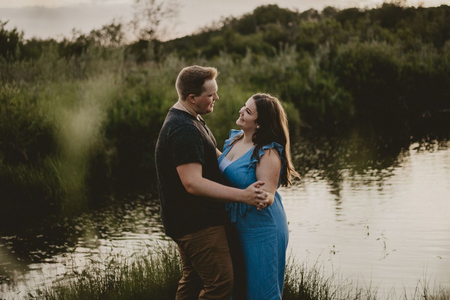 Romantic couple in casual attire embracing near a peaceful lake at sunset.