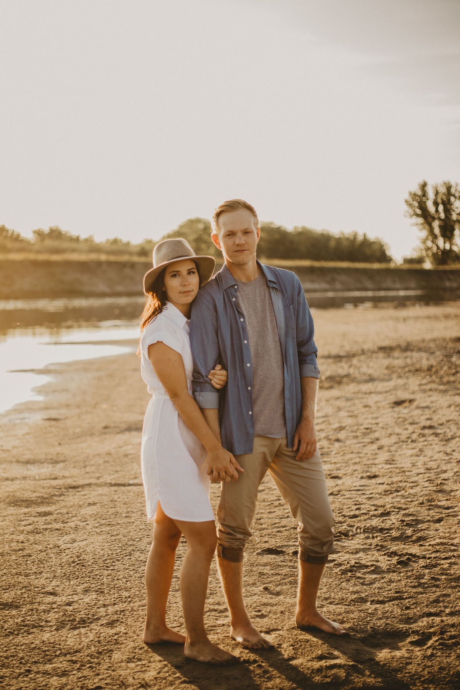 A couple poses together on a beach during golden hour wearing coordinated casual outfits.