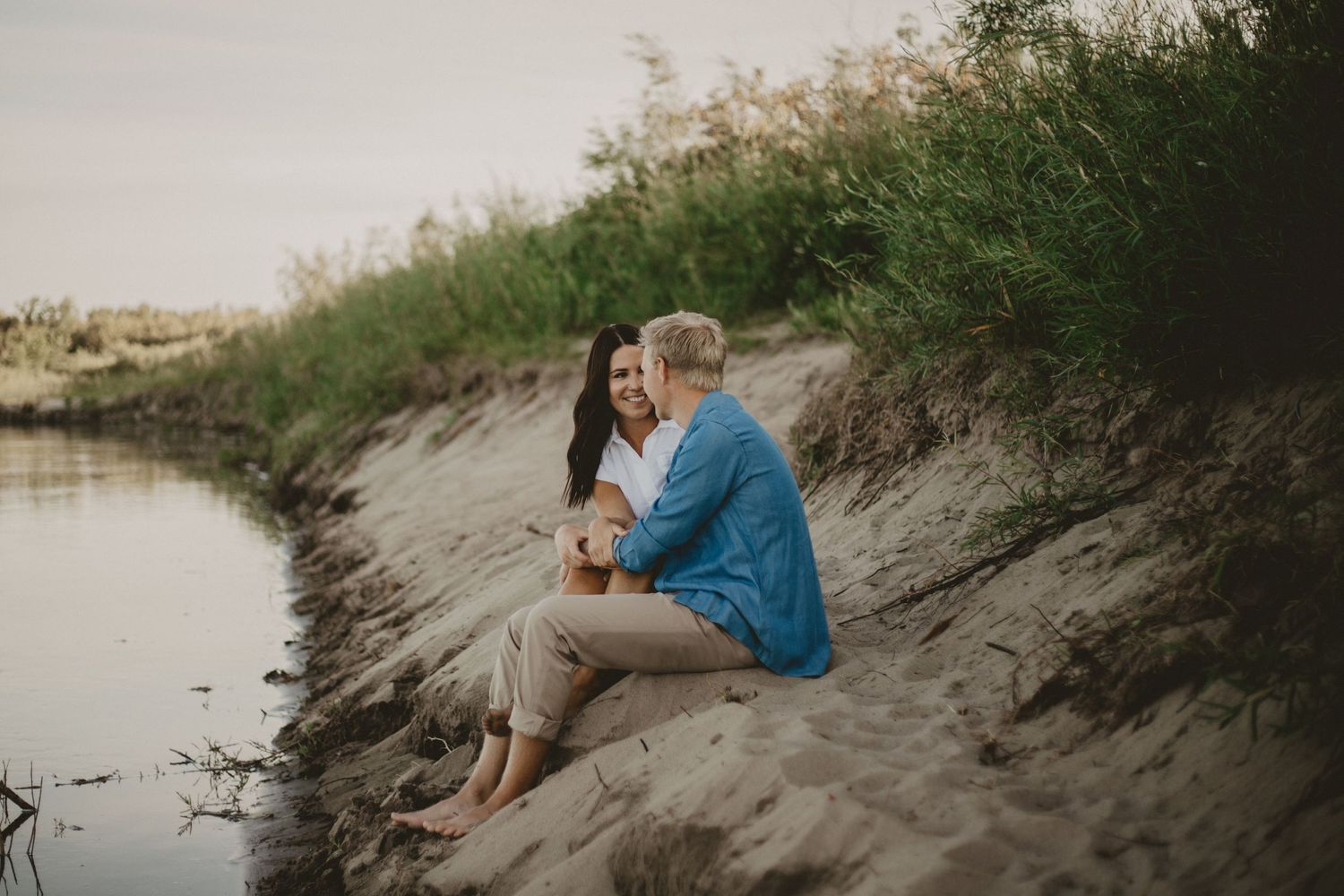 Tender moment between couple sitting on riverbank during golden hour.