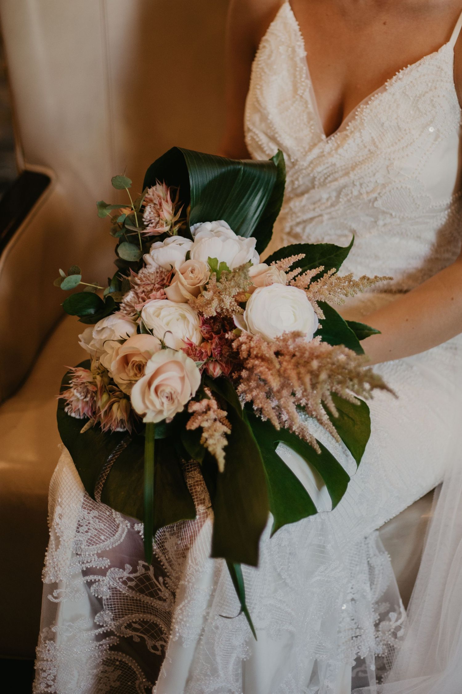 Elegant bridal bouquet featuring blush roses, tropical leaves and pampas grass against a white lace wedding dress.