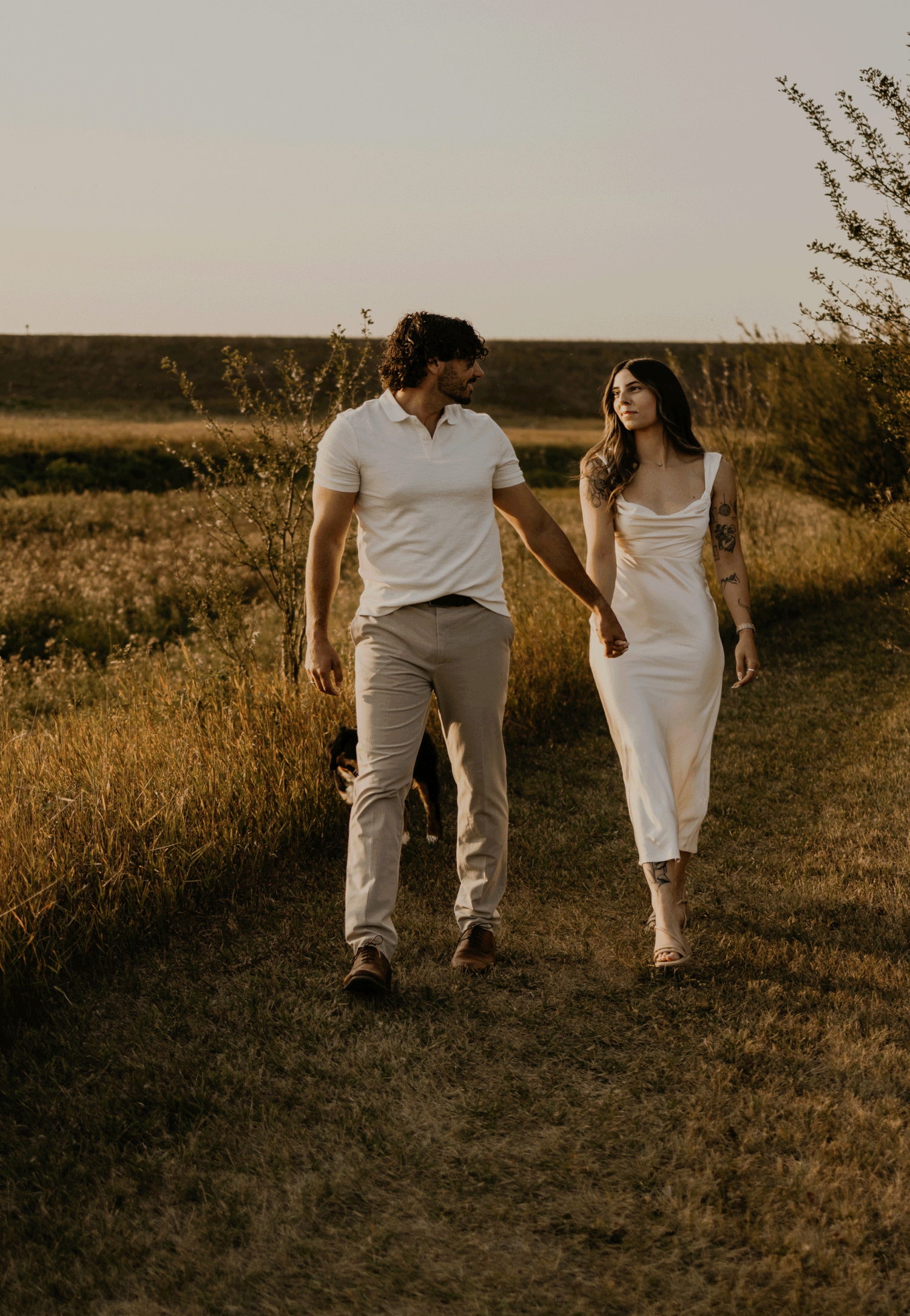 Couple in white outfits walking together through golden grass field during sunset.