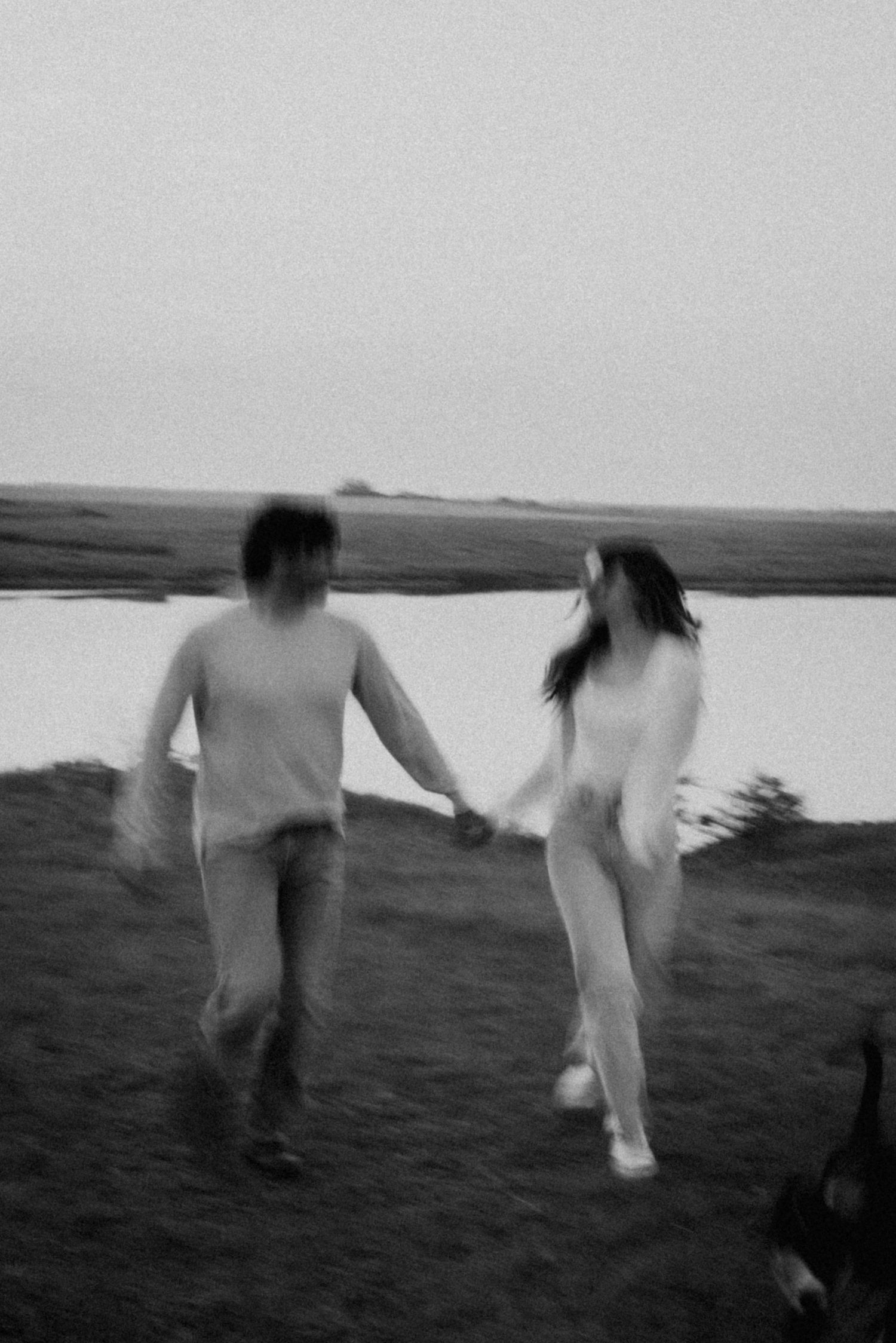 A blurry black and white photo of a couple running hand in hand by a lake.