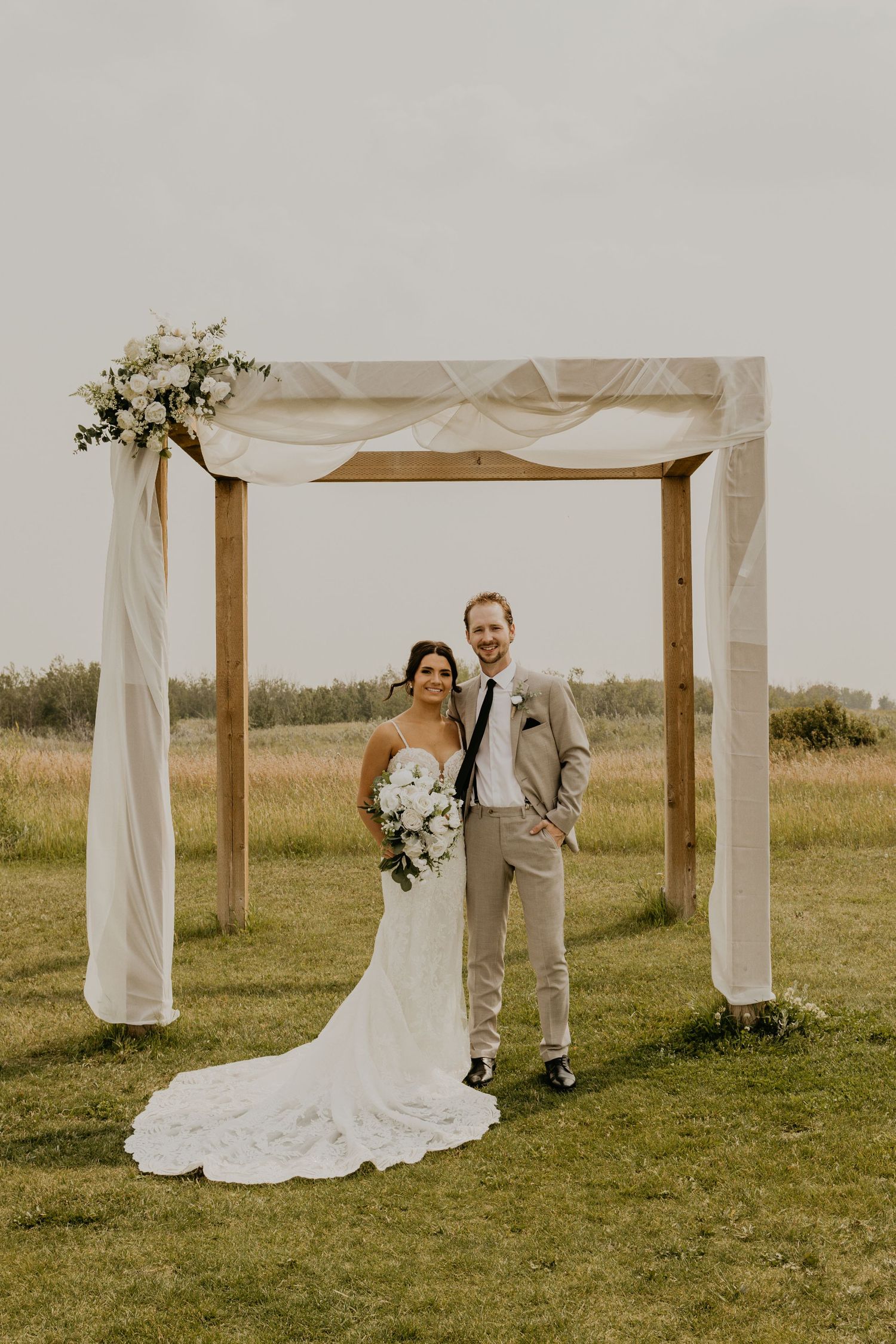 Newlyweds pose under draped wooden wedding arch in outdoor field setting.
