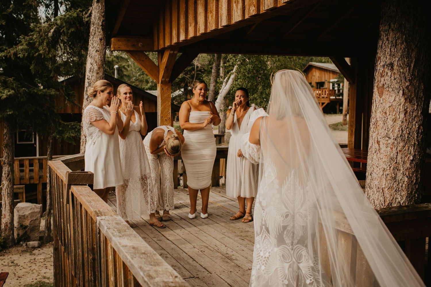 Bridesmaids in light colored dresses react emotionally on a wooden deck during a wedding ceremony.