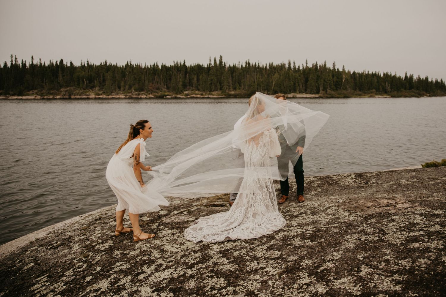 Wedding dress flowing in wind on rocky shoreline with lake view.
