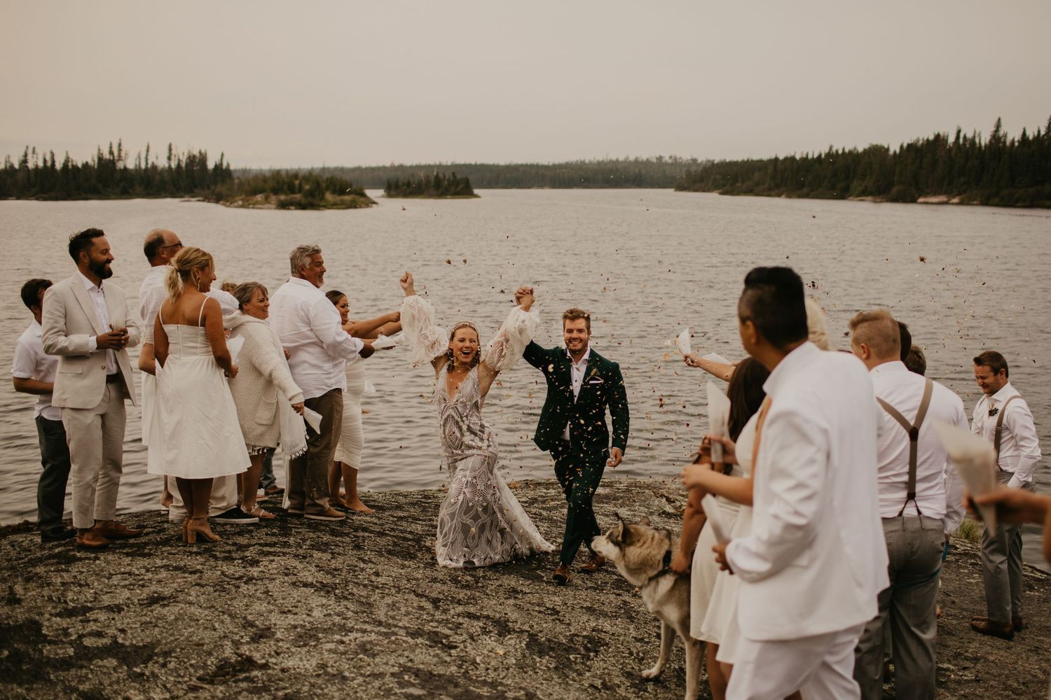 Wedding celebration with wedding party in white attire cheering by a lakeside.