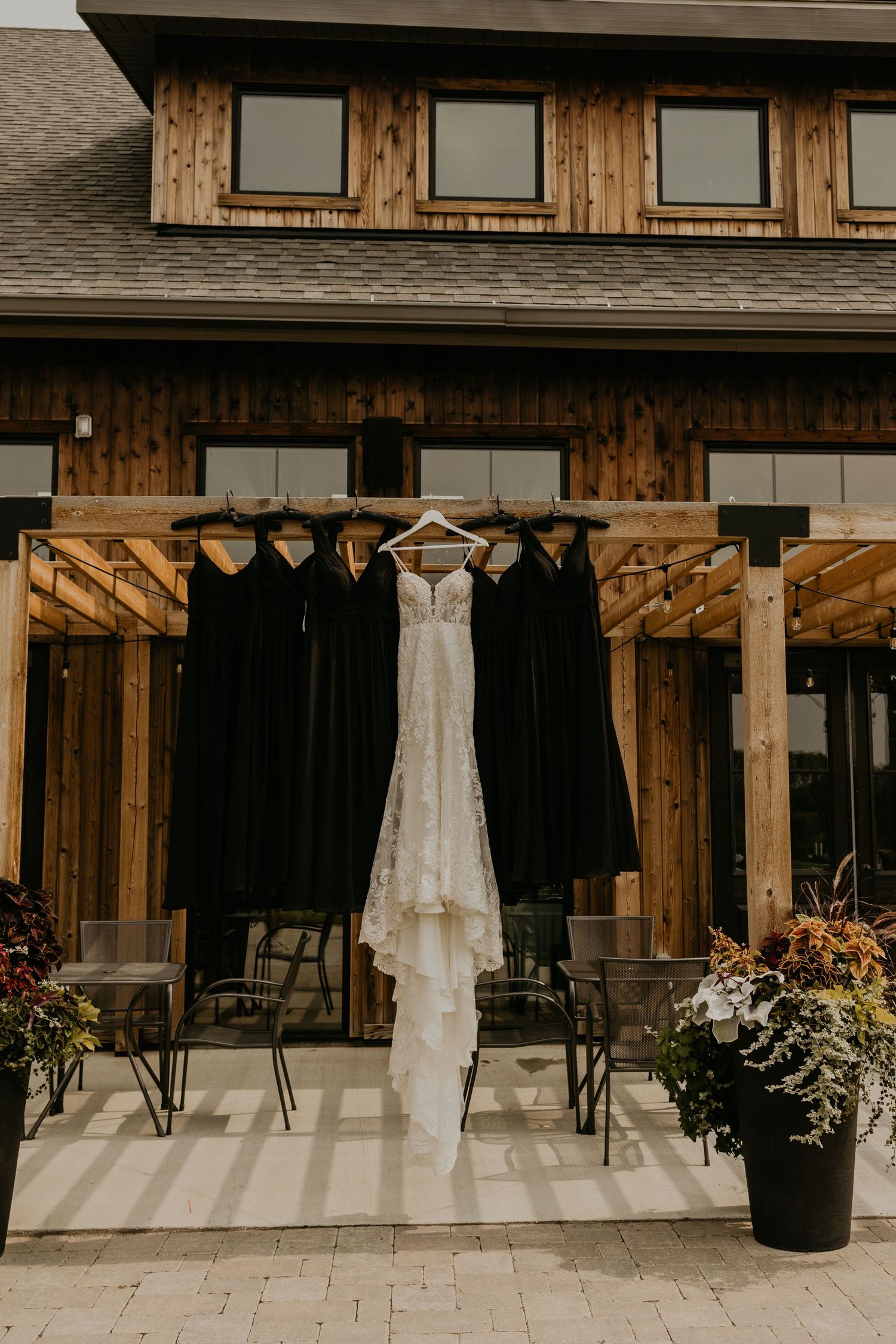 White wedding dress and black bridesmaid dresses hanging on rustic wooden porch with potted flowers.