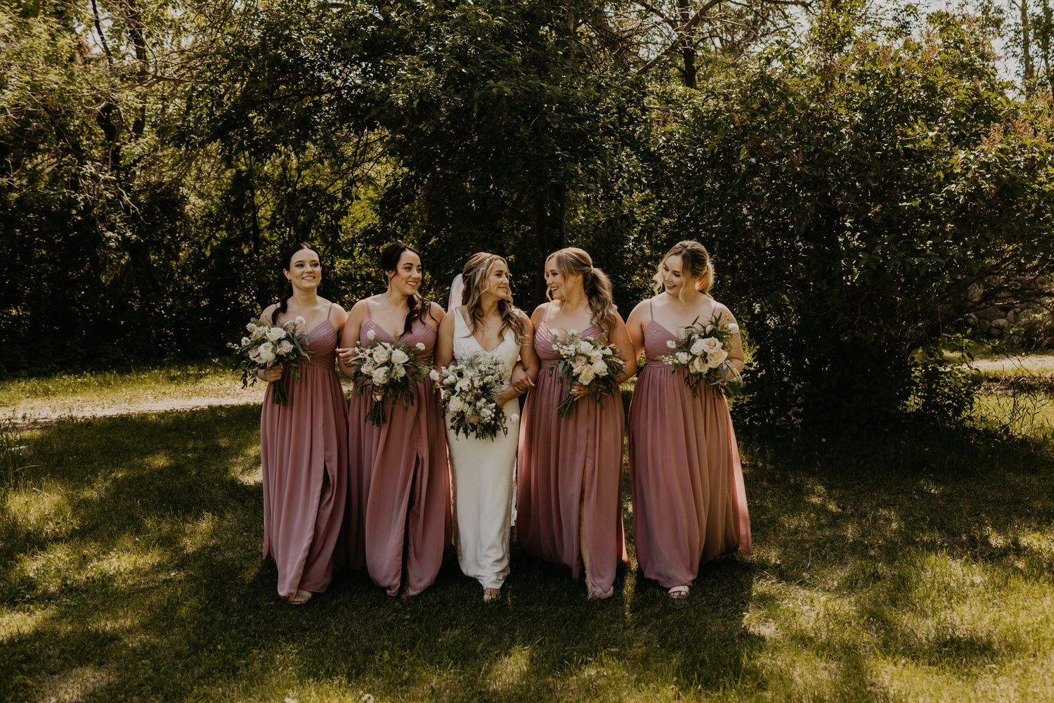 Bridal party in rose-colored dresses stands together holding white bouquets in outdoor setting.