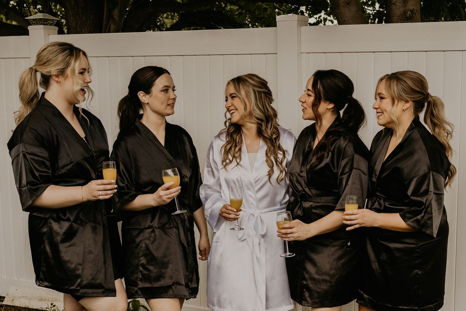 Wedding party wearing matching black robes and one white robe toasting with champagne glasses outdoors.