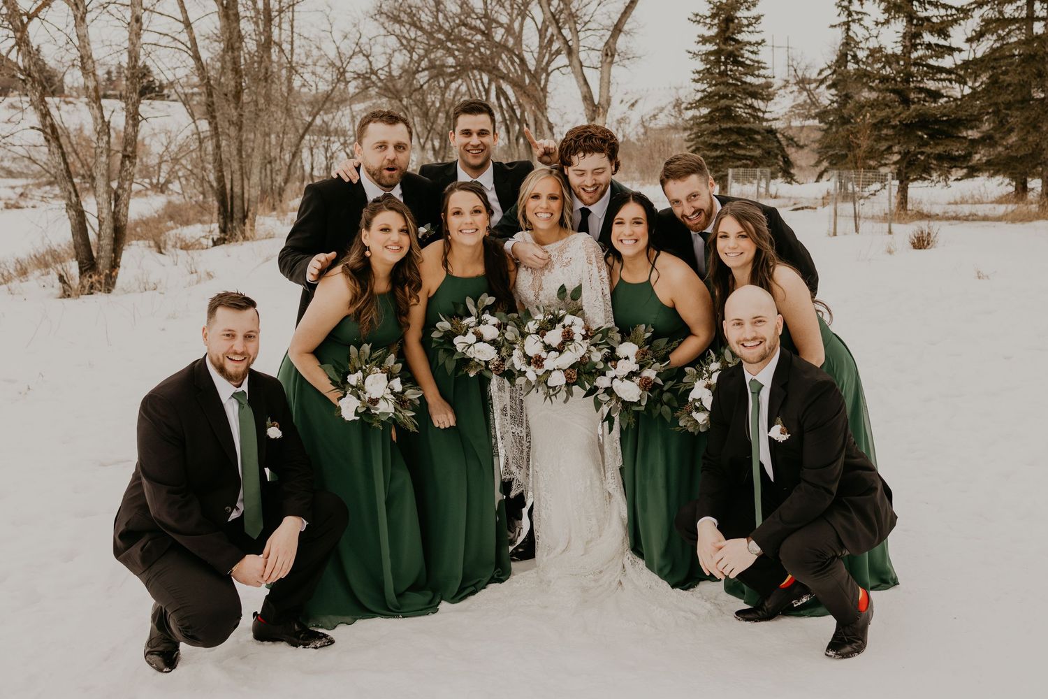 Wedding party in black suits and emerald green dresses poses casually in the snow.