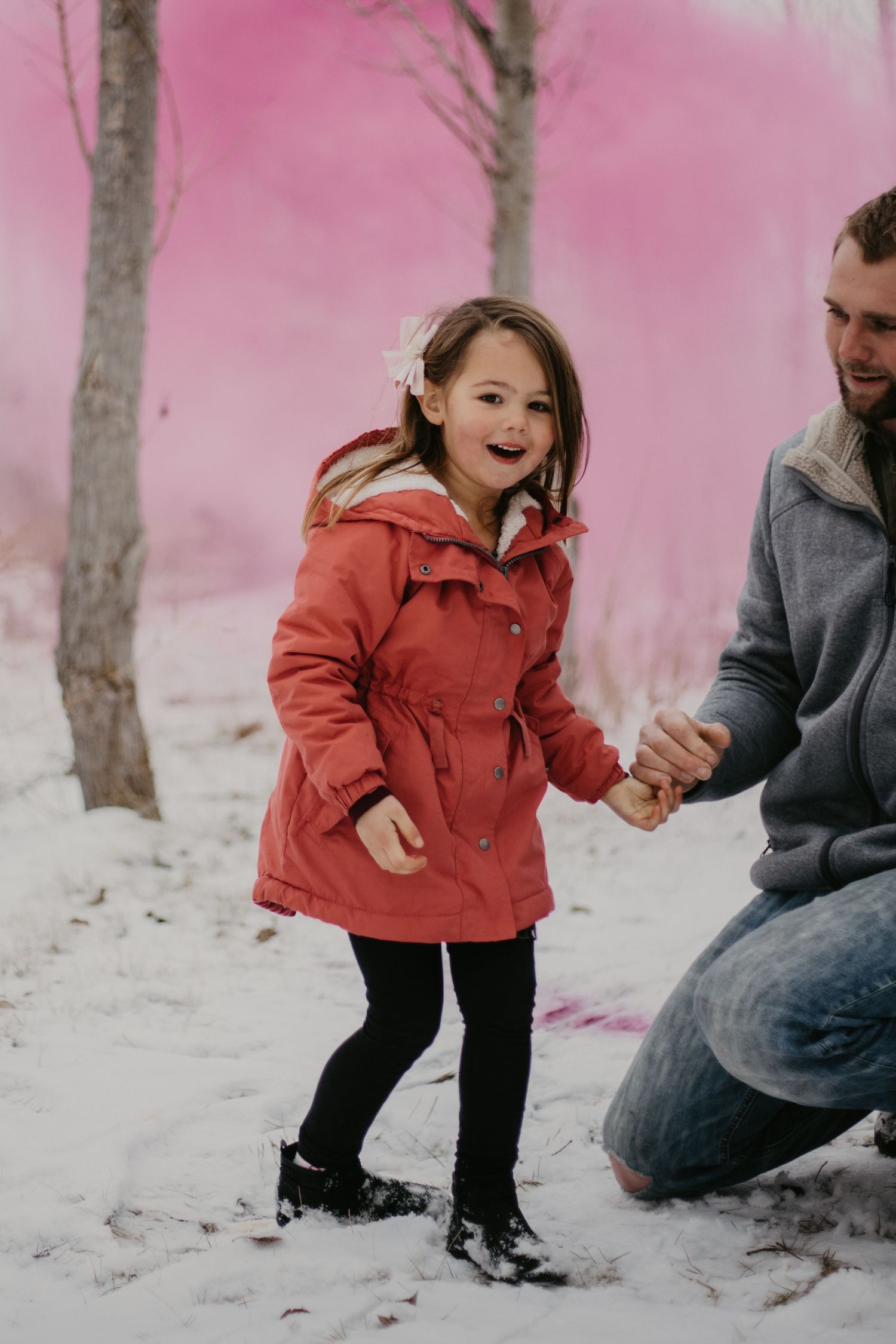 A joyful child wearing a red winter coat plays in the snow while holding someone's hand on a pink-tinted winter day.