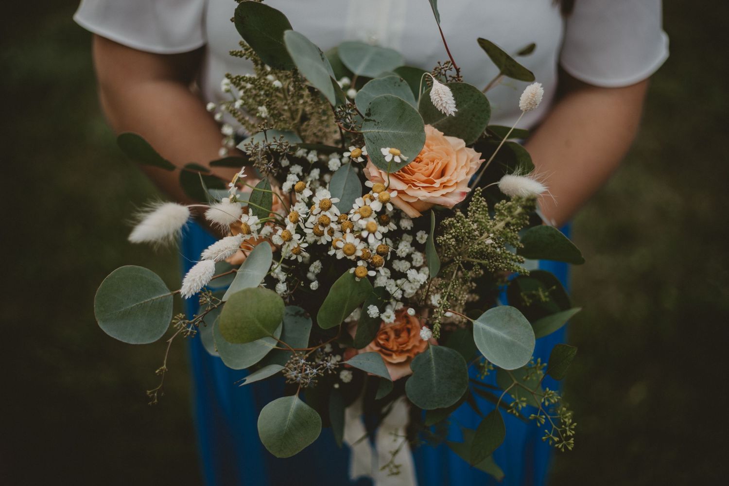 A bouquet of peach roses, baby's breath and eucalyptus leaves held against blue jeans.