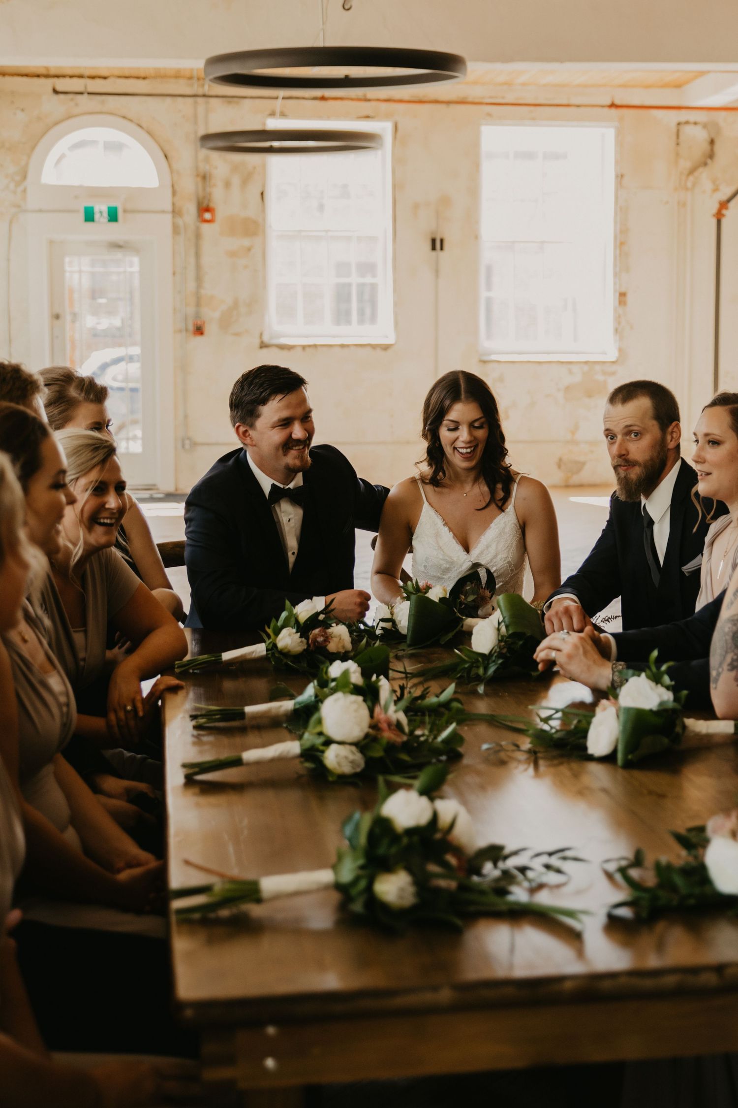 Wedding reception scene with guests gathered around a rustic wooden table decorated with floral arrangements.