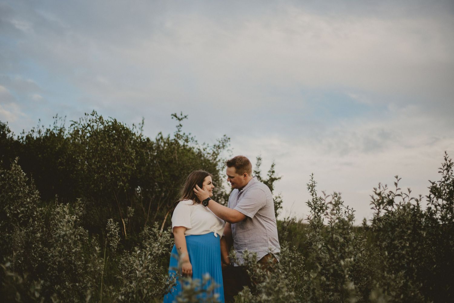 A couple laughs together while standing in a natural meadow setting.
