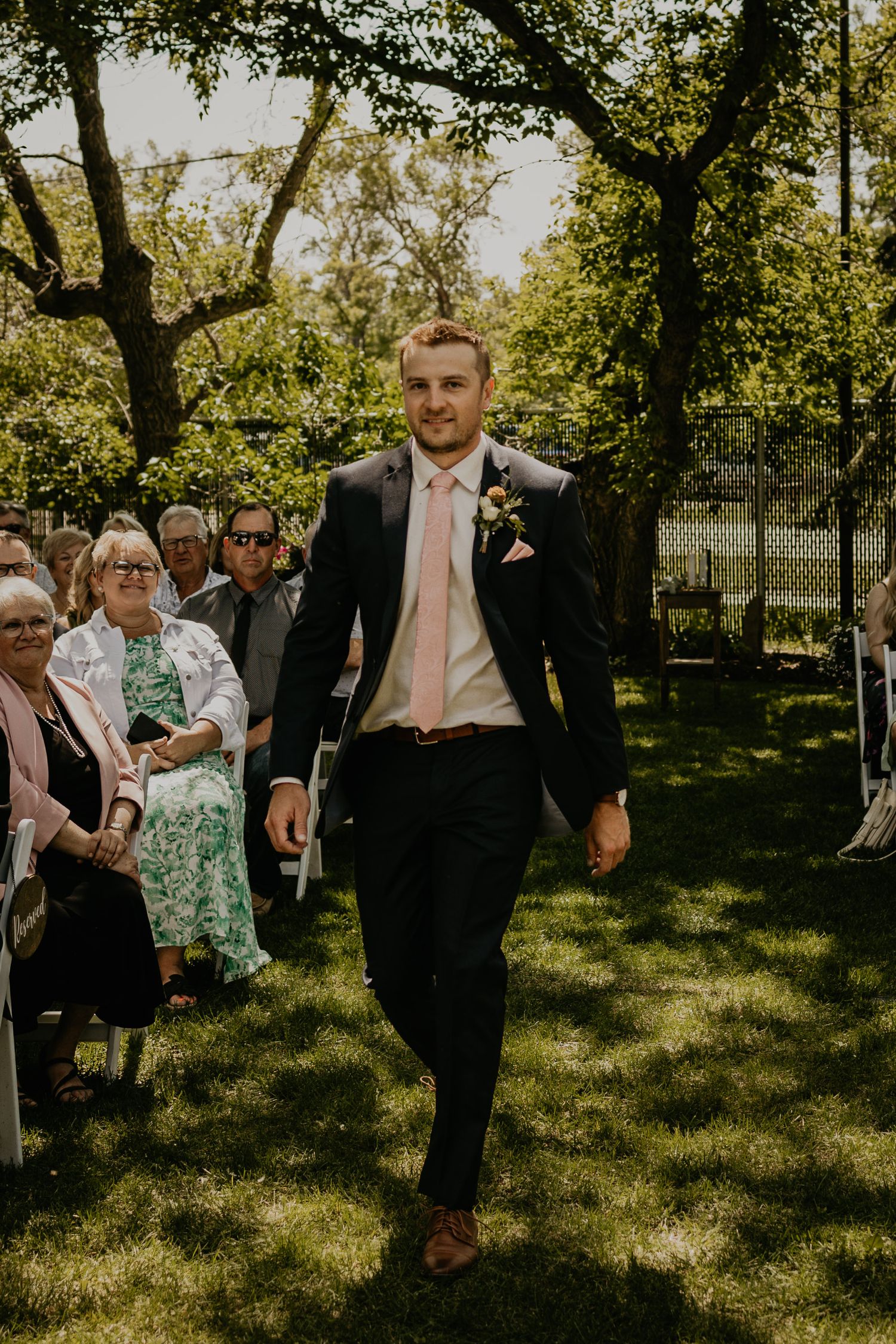 Groom walks down outdoor ceremony aisle in black suit with pink tie.