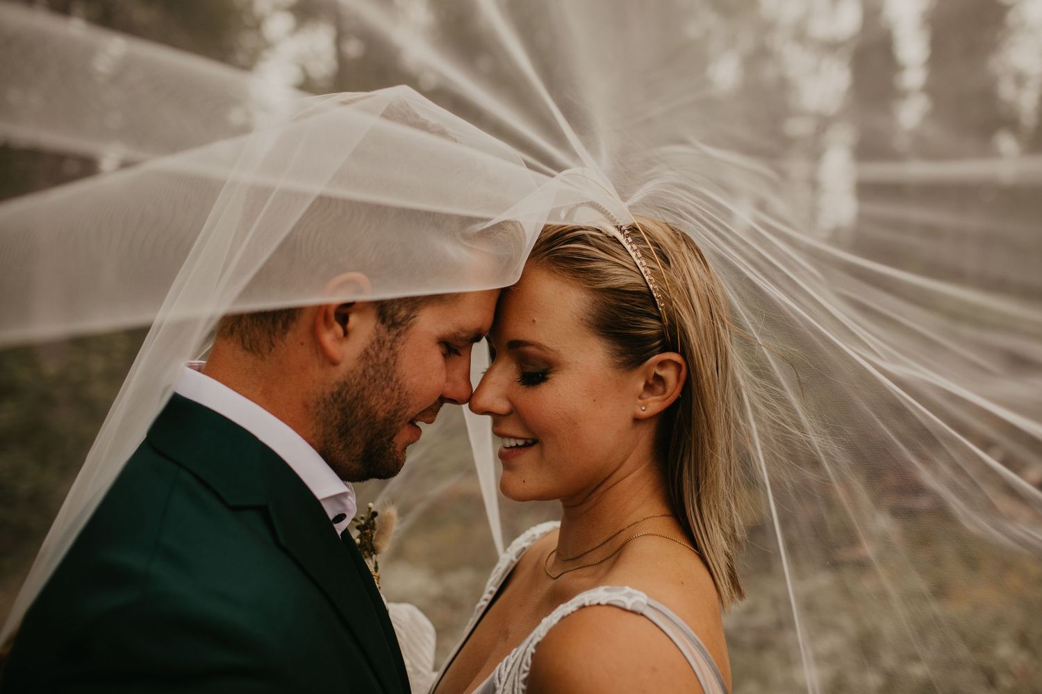 Intimate wedding moment captured in a forest setting with a flowing veil and emerald green suit.