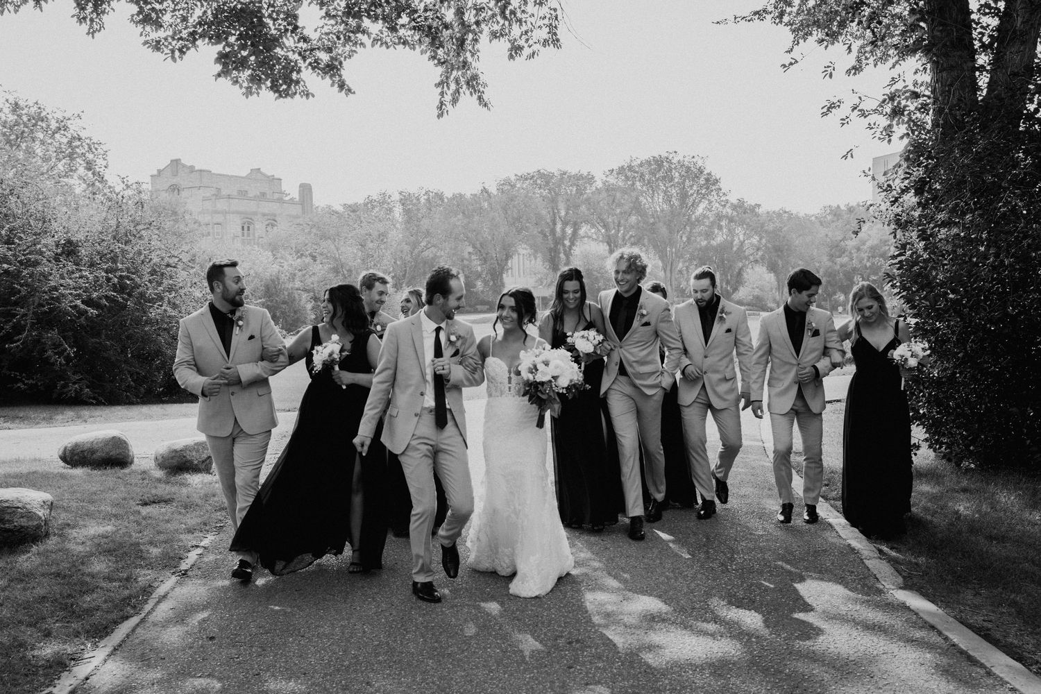 Wedding party in black dresses and beige suits posing together on outdoor pathway.